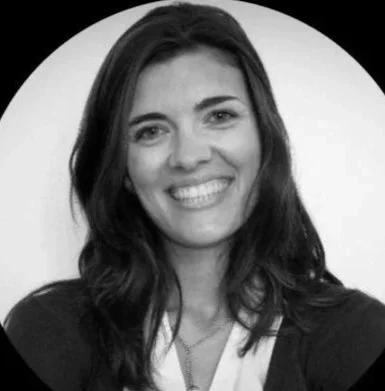 Une femme souriante avec des cheveux longs et bruns, portant un blazer noir et une chemise blanche.