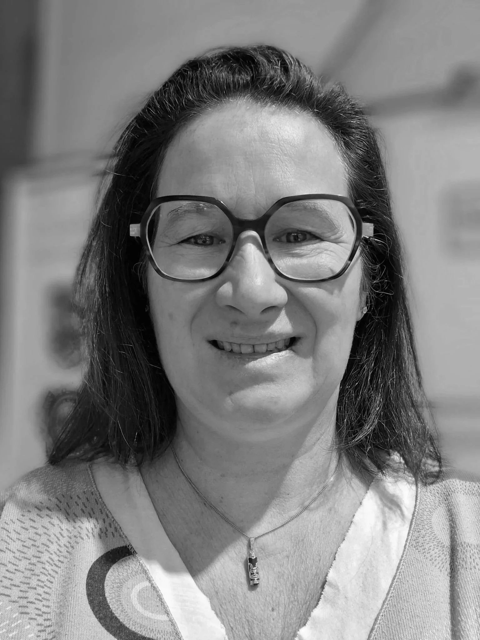 Une femme souriante porte des lunettes et un collier avec un pendentif, photo en noir et blanc.
