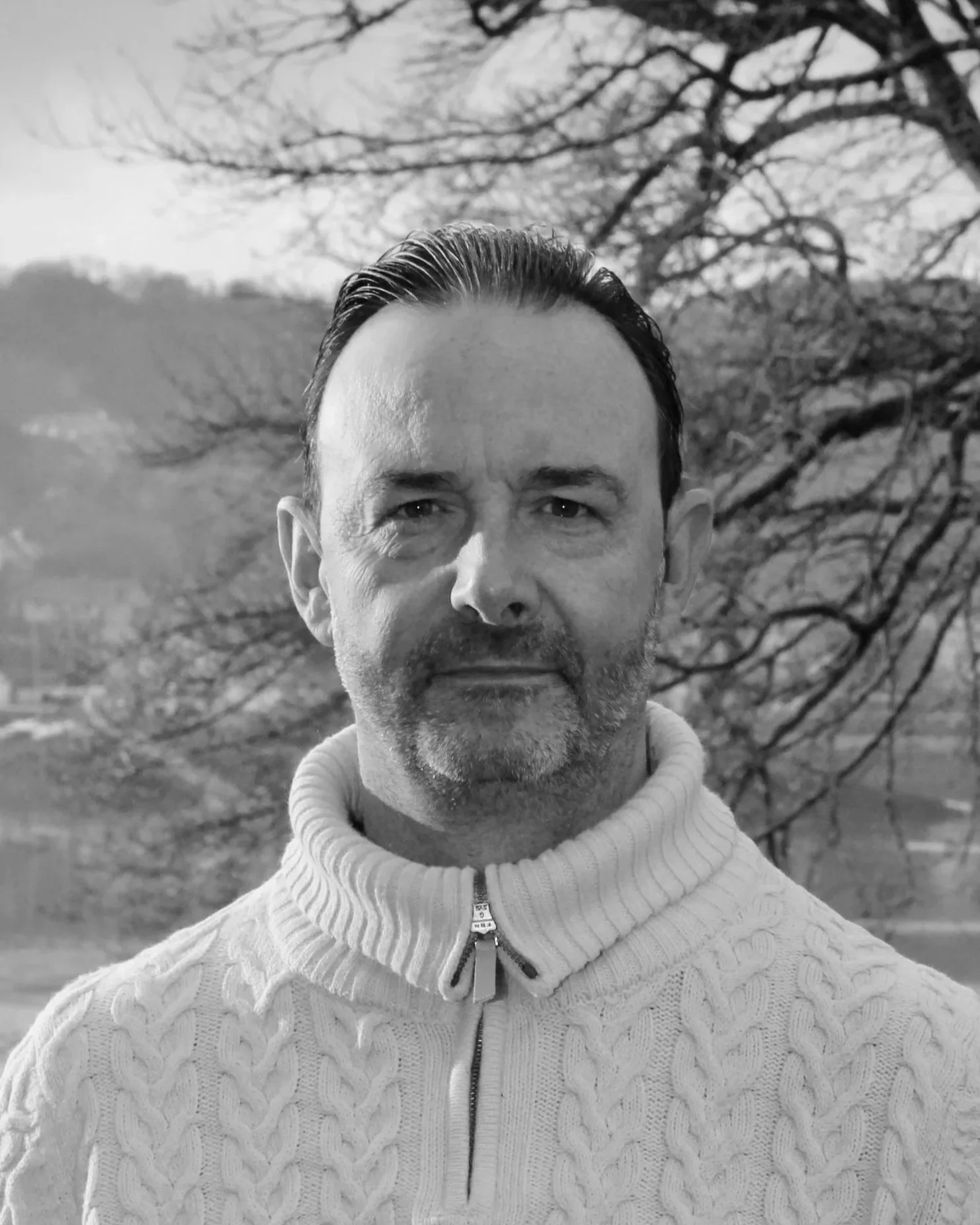 Un homme avec des cheveux courts et une barbe, portant un pullover à col roulé en cachemire avec une fermeture éclair, se tenant devant un paysage de campagne avec des arbres sans feuilles et des collines en arrière-plan, en noir et blanc.