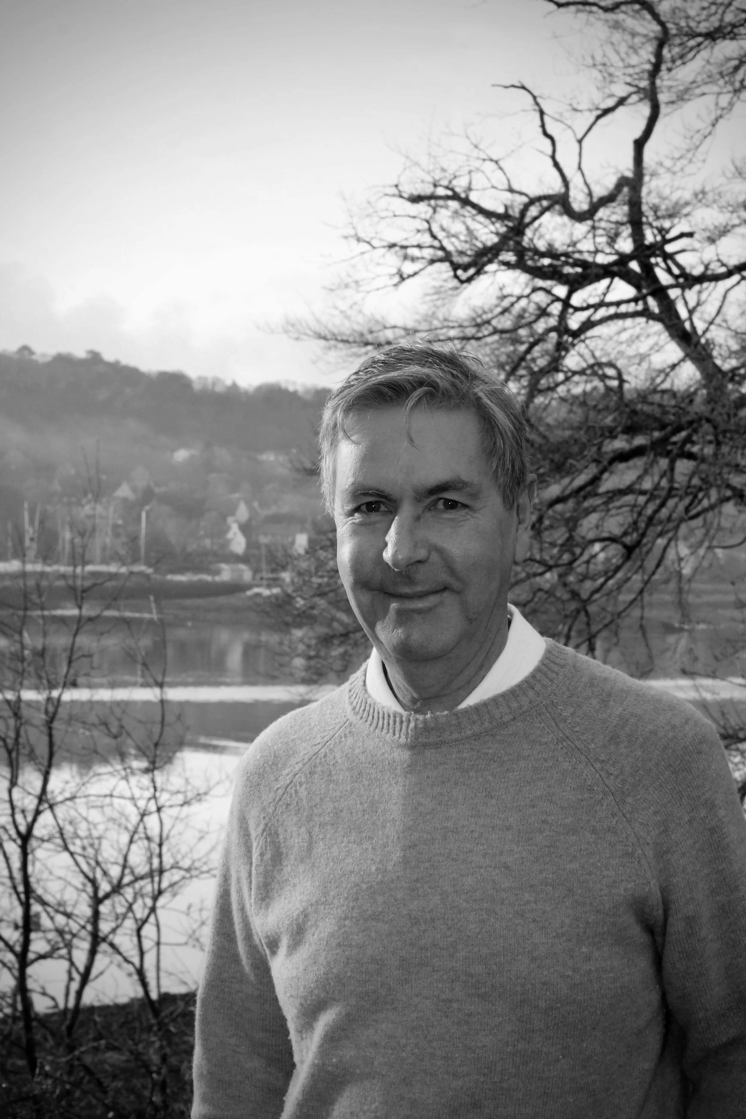 Hommes souriant en plein air, avec un arbre sans feuilles et un lac en arrière-plan, en noir et blanc.