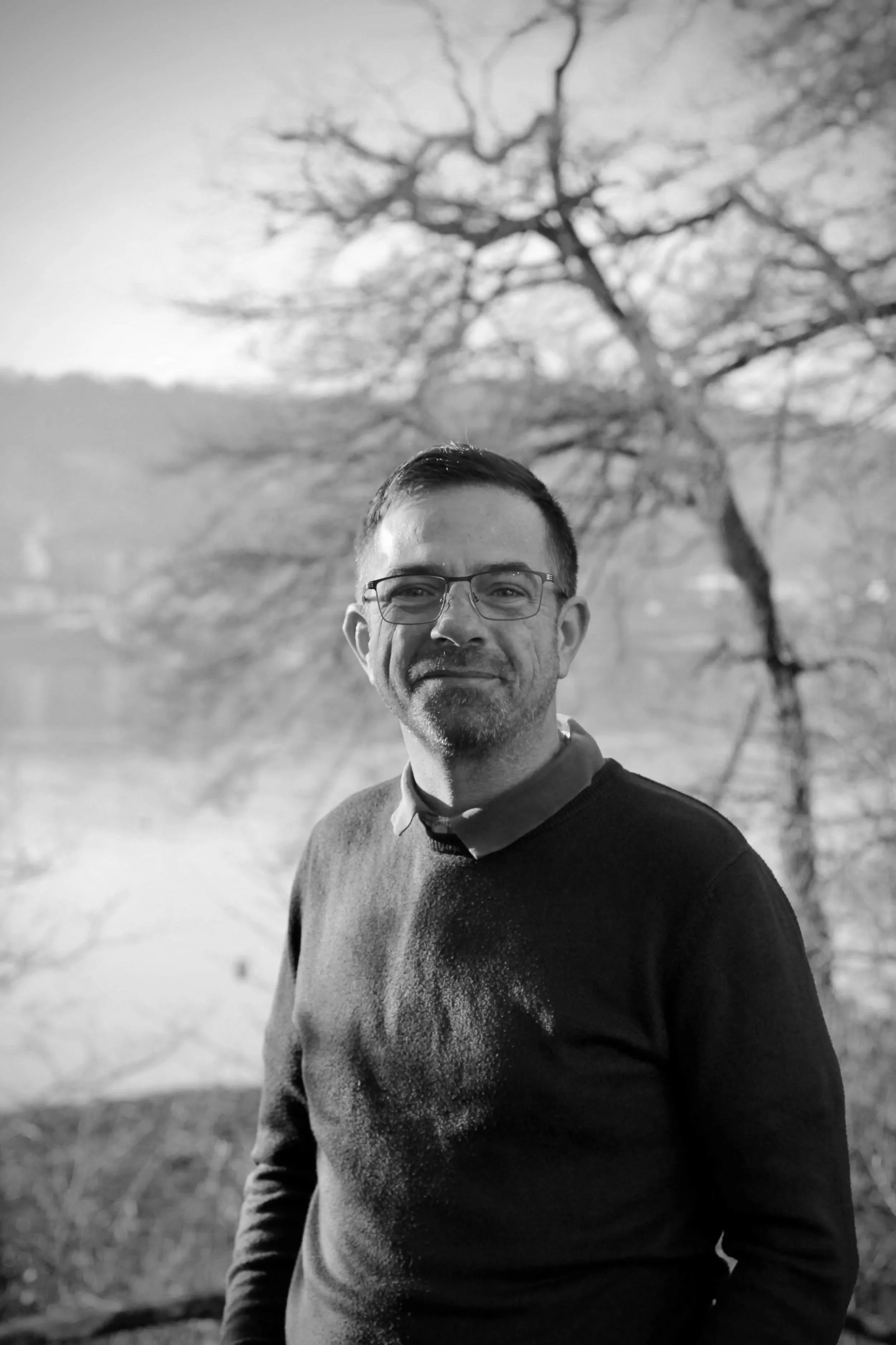 Un homme souriant portant des lunettes, debout à l'extérieur avec un arbre sans feuilles en arrière-plan en noir et blanc.
