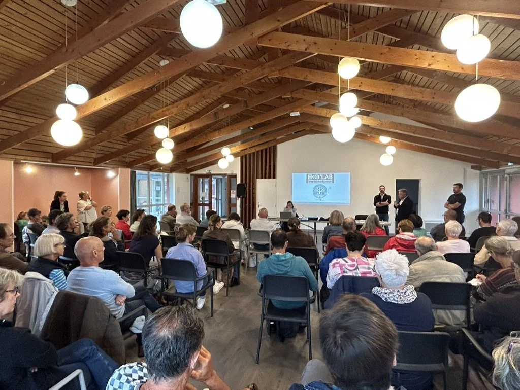 Une salle de conférence pleine de personnes assises, écoutant des intervenants devant un écran avec le logo 'EKO LAB'. Il y a des luminaires suspendus en forme de globes en bois au plafond en bois apparent.