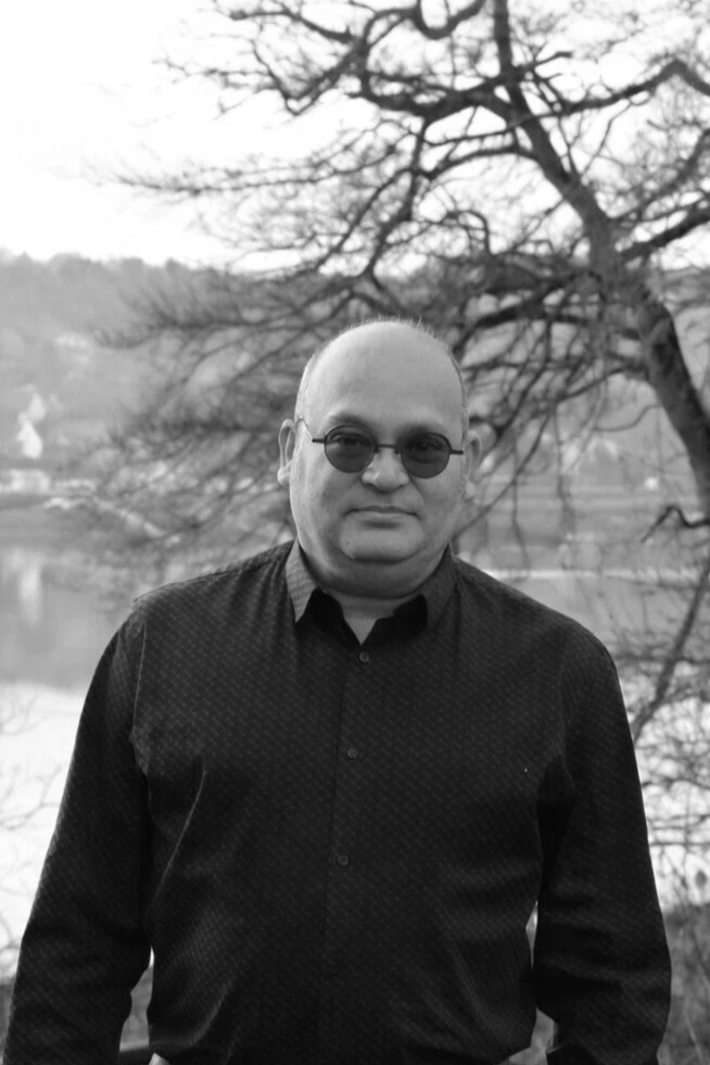 Un homme portant des lunettes noires, habillé d'une chemise noire, se tient devant un arbre nu et un paysage flou avec une rivière, en noir et blanc.