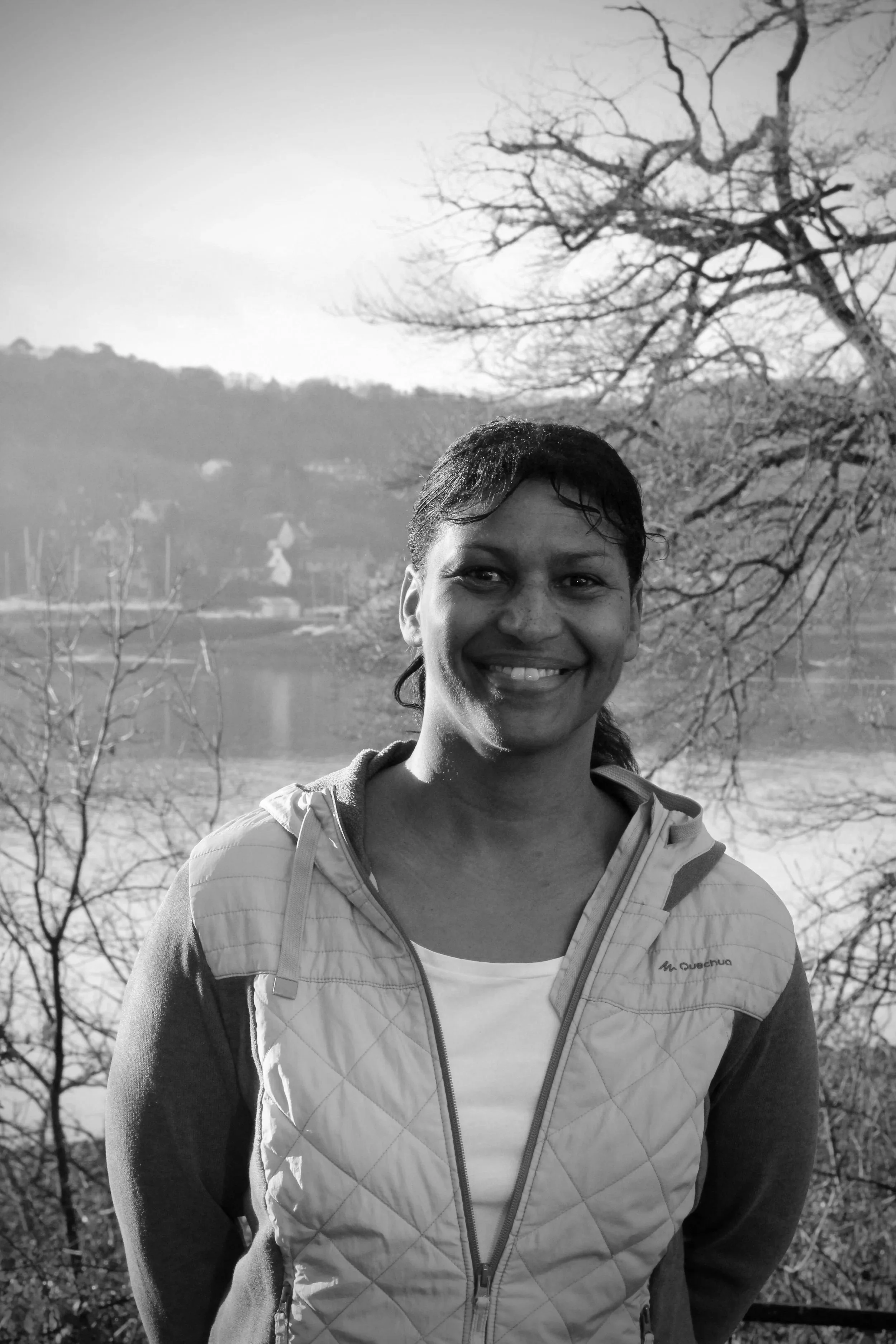 Une femme souriante avec cheveux courts, portant une veste, se tenant devant un paysage naturel comprenant un lac, des montagnes, et des arbres sans feuilles, en noir et blanc.