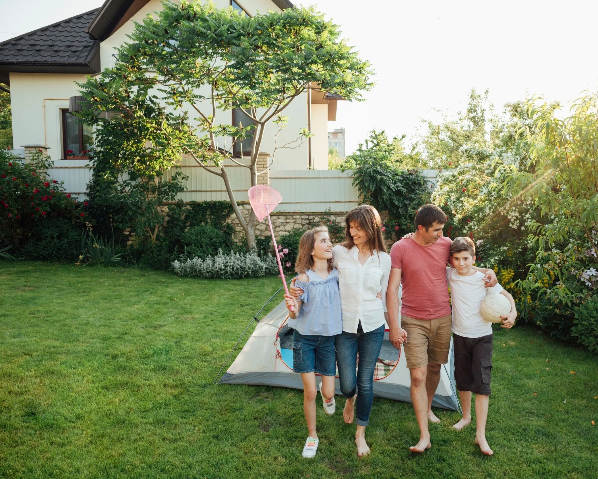Famille de quatre personnes marchant dans leur jardin avec une tente en arrière-plan. La mère, le père, une fille et un garçon, tous souriants. La fille tient une petite épuisette rose, le garçon tient un ballon de basket, et le père porte un ballon de football. Il fait beau et ensoleillé.