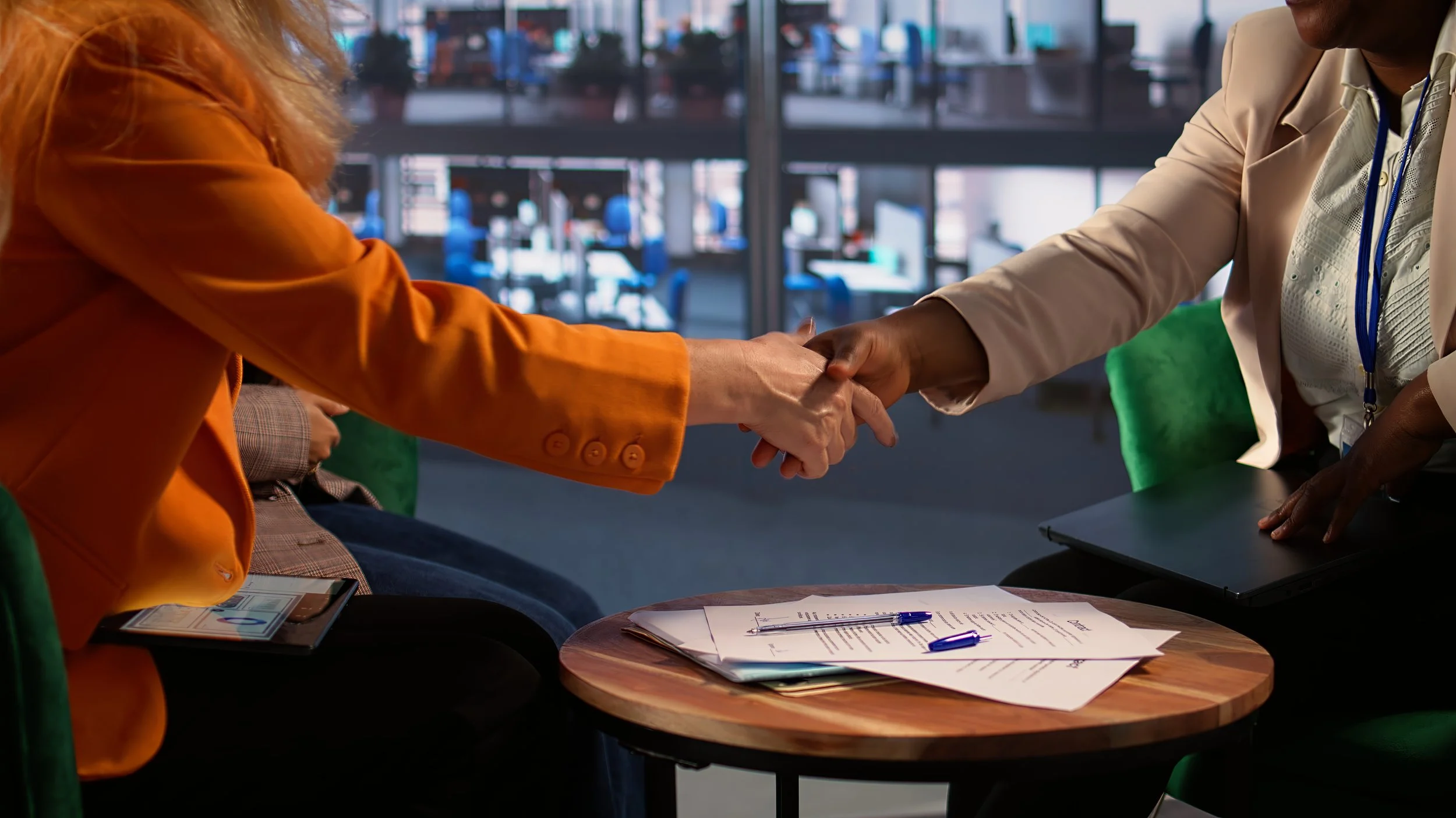 Deux personnes se serrent la main lors d'une rencontre professionnelle, avec des documents et des stylos sur une petite table en bois, dans un environnement moderne de bureau.