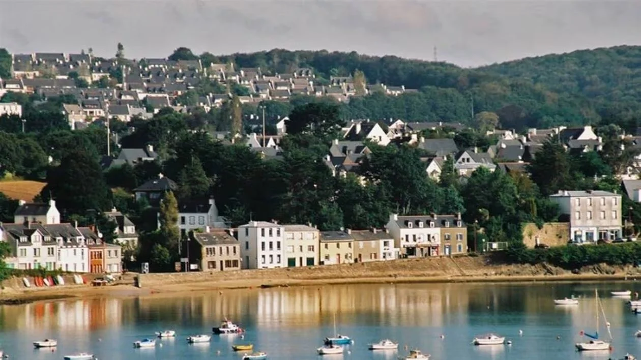 Village côtier avec maisons colorées et bateaux sur l'eau, entouré de collines verdoyantes.