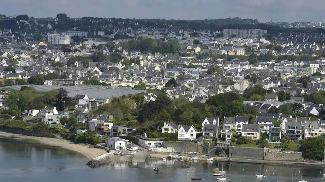 Une vue panoramique d'une ville côtière avec de nombreux bâtiments résidentiels blanches et grises, une plage au premier plan, et un petit port avec des bateaux sur l'eau.