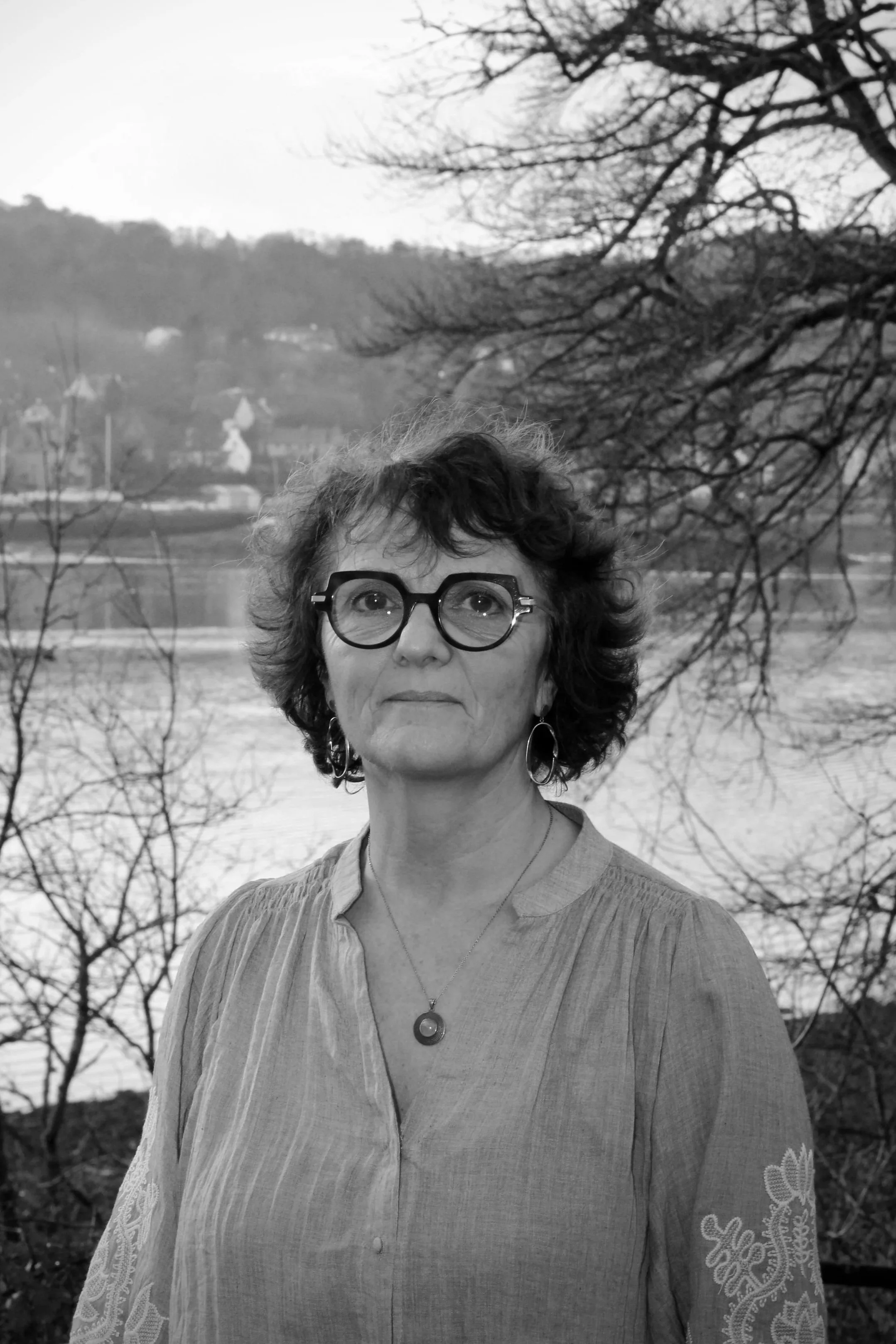 Portrait en noir et blanc d'une femme portant des lunettes rondes et un pendentif, avec un arrière-plan d'arbres et d'une rivière.