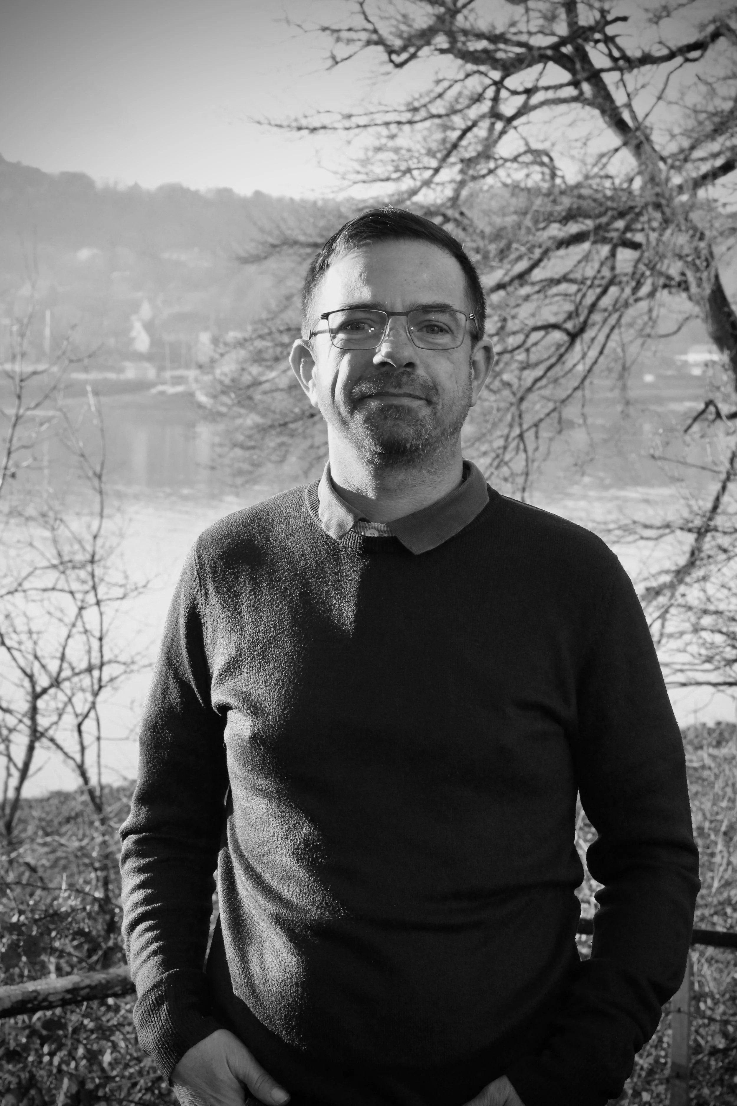 Un homme portant des lunettes et un pull sombre, debout près d'un arbre avec un paysage de rivière et de montagnes en arrière-plan, photographié en noir et blanc.