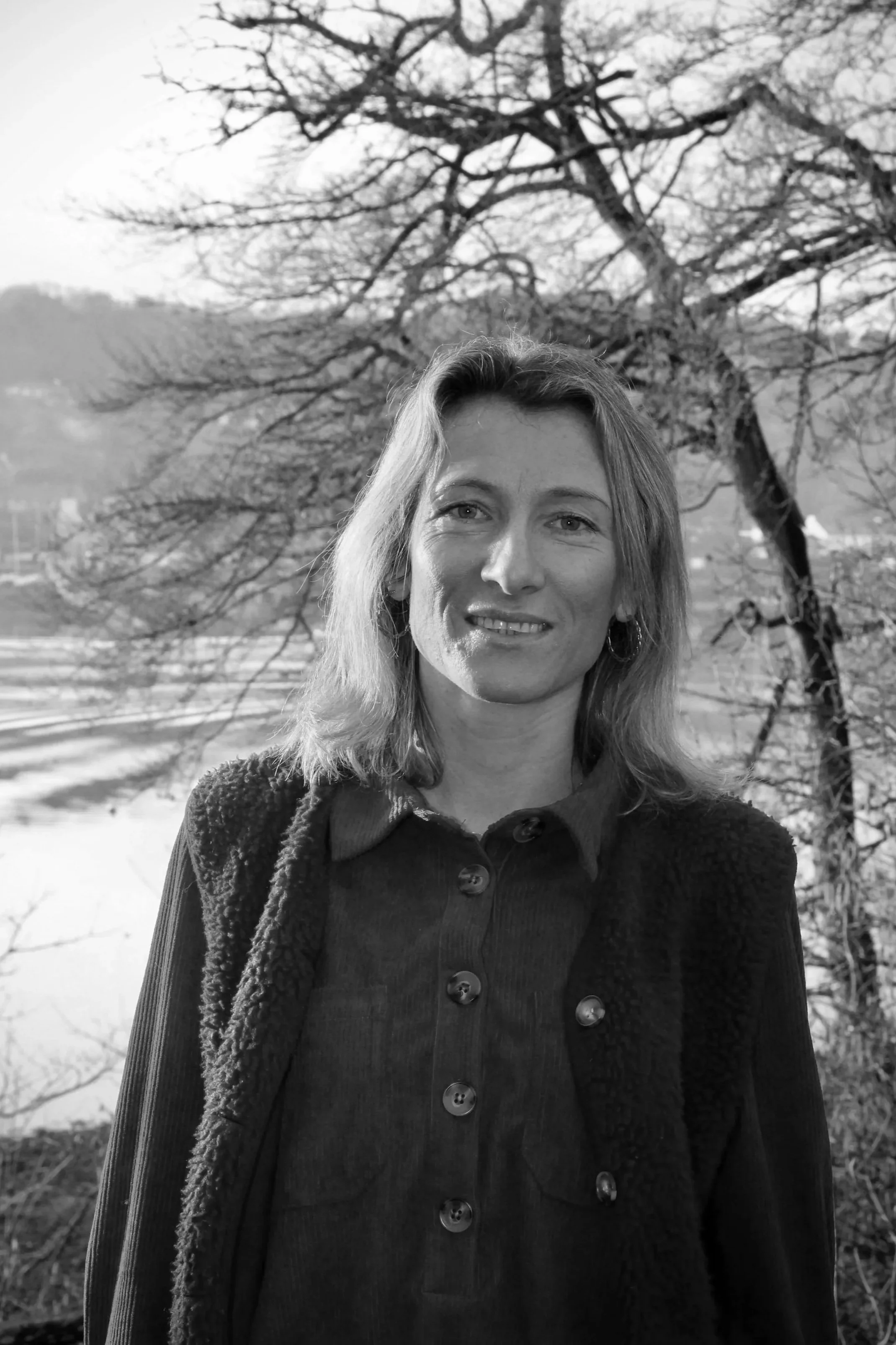 Une femme avec des cheveux mi-longs, souriante, en pantalon et veste en laine, se tient devant un arbre nu avec un paysage de montagnes et de champs au loin, en arrière-plan.