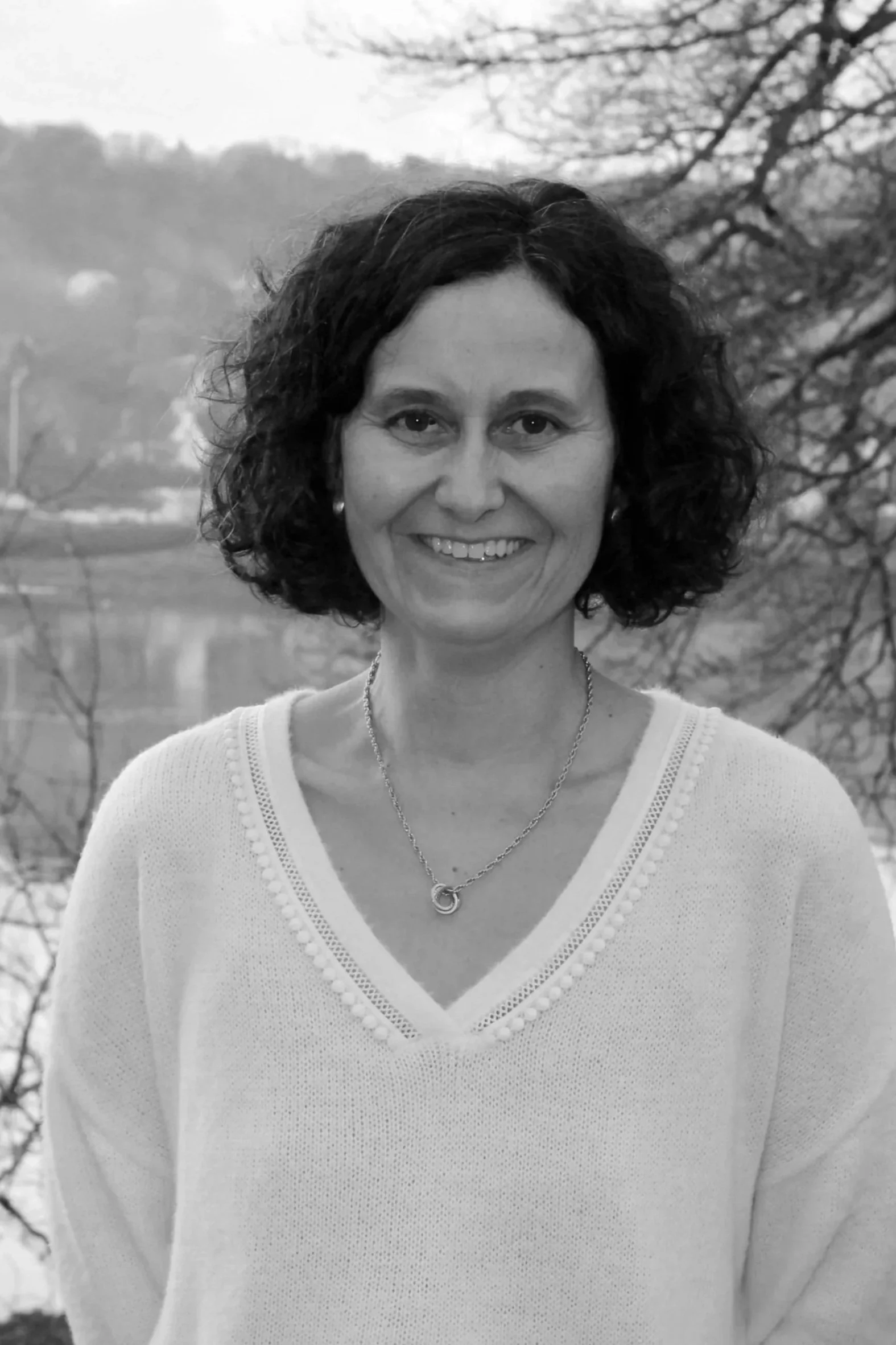 Une femme souriante avec des cheveux bouclés, portant un pull blanc avec un collier autour du cou, en extérieur avec des arbres et un corps d'eau en arrière-plan.