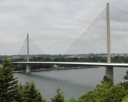 Pont suspendu au-dessus d'une rivière, avec paysage urbain en arrière-plan et arbres verts au premier plan.