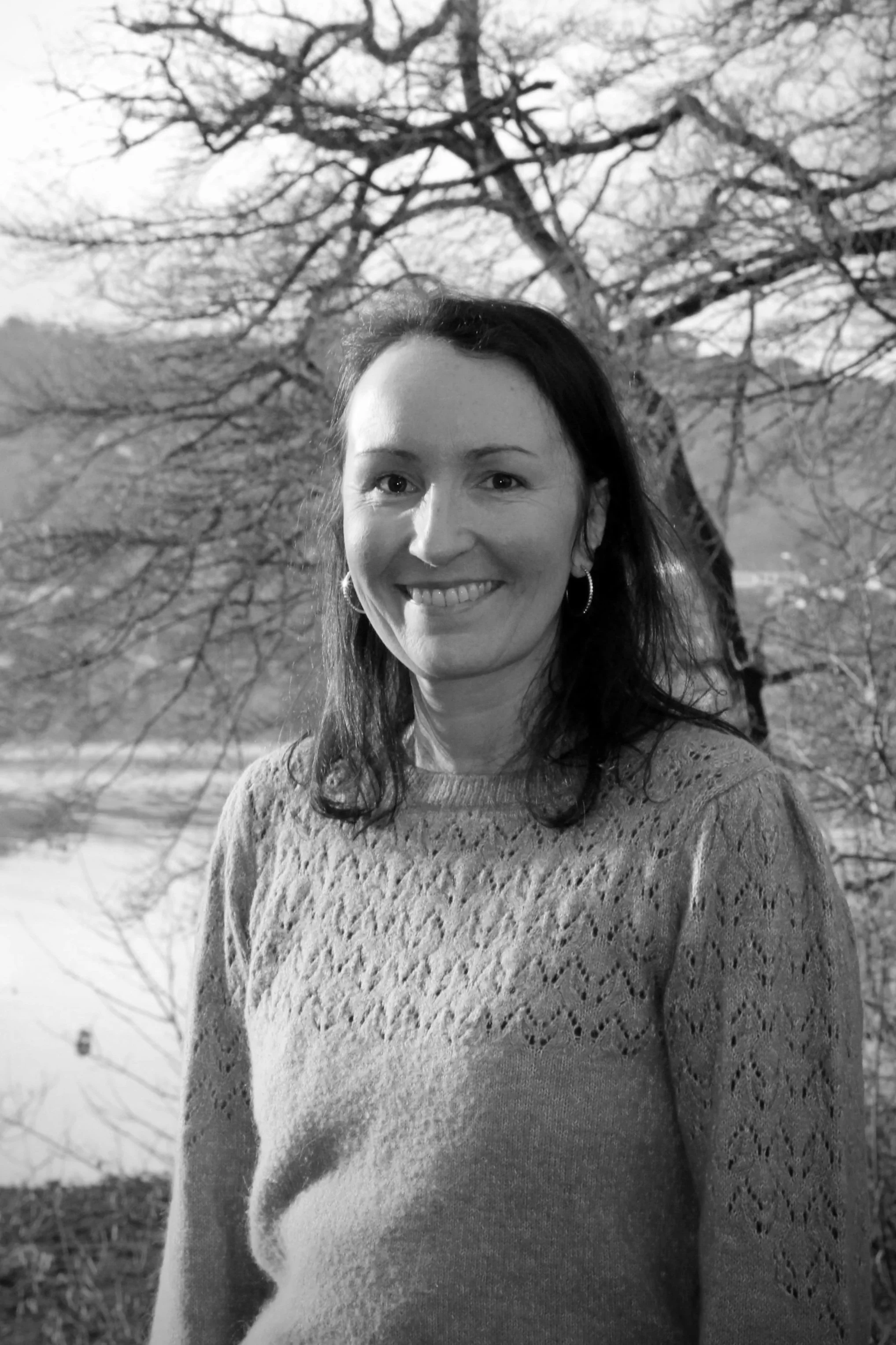 Portrait d'une femme souriante devant un arbre nu en extérieur, en noir et blanc.