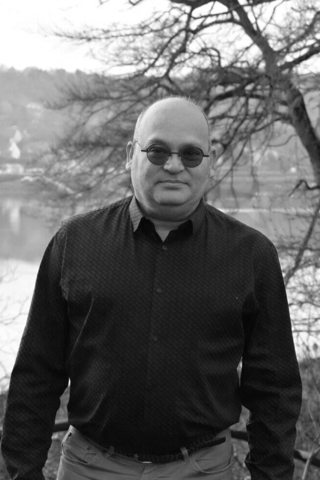 Un homme vêtu d'une chemise noire à motifs, portant des lunettes de soleil, se tenant devant un arbre sans feuilles et un lac.