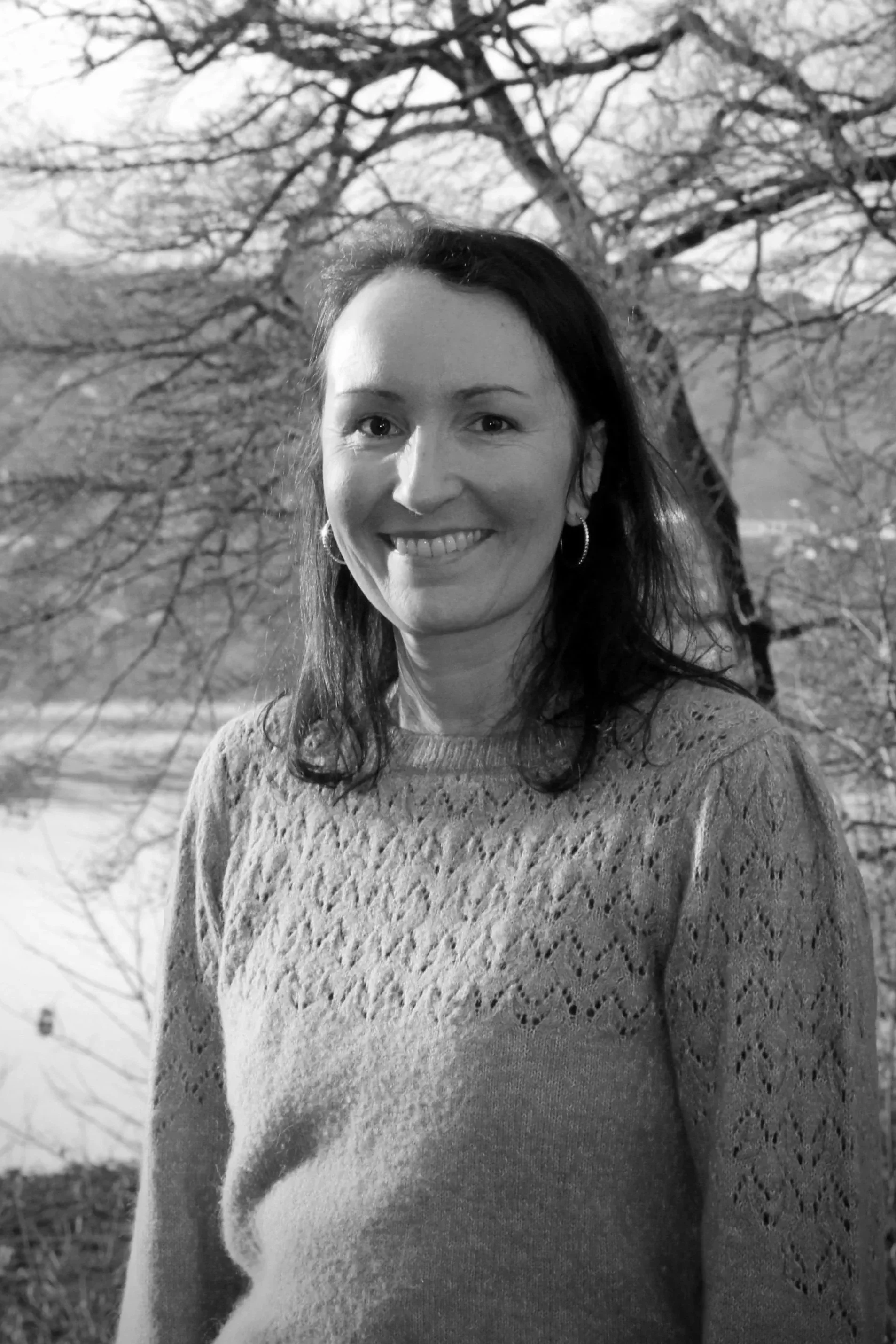Une femme souriante avec des cheveux longs foncés portait un pull en tricot, avec un arbre en arrière-plan en extérieur.