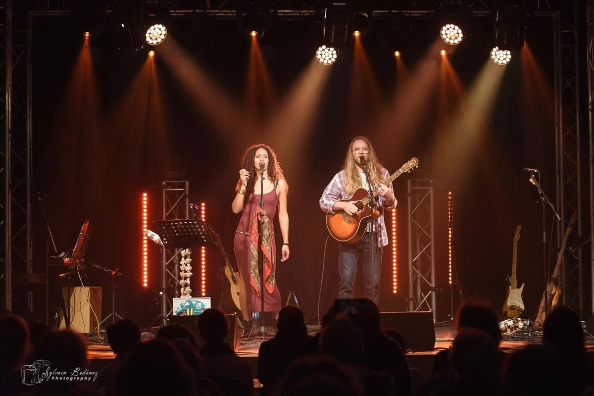 Deux musiciens sur scène, une femme chante avec un micro et un homme joue de la guitare au piano, éclairés par des spots lumineux, un public assis en avant.