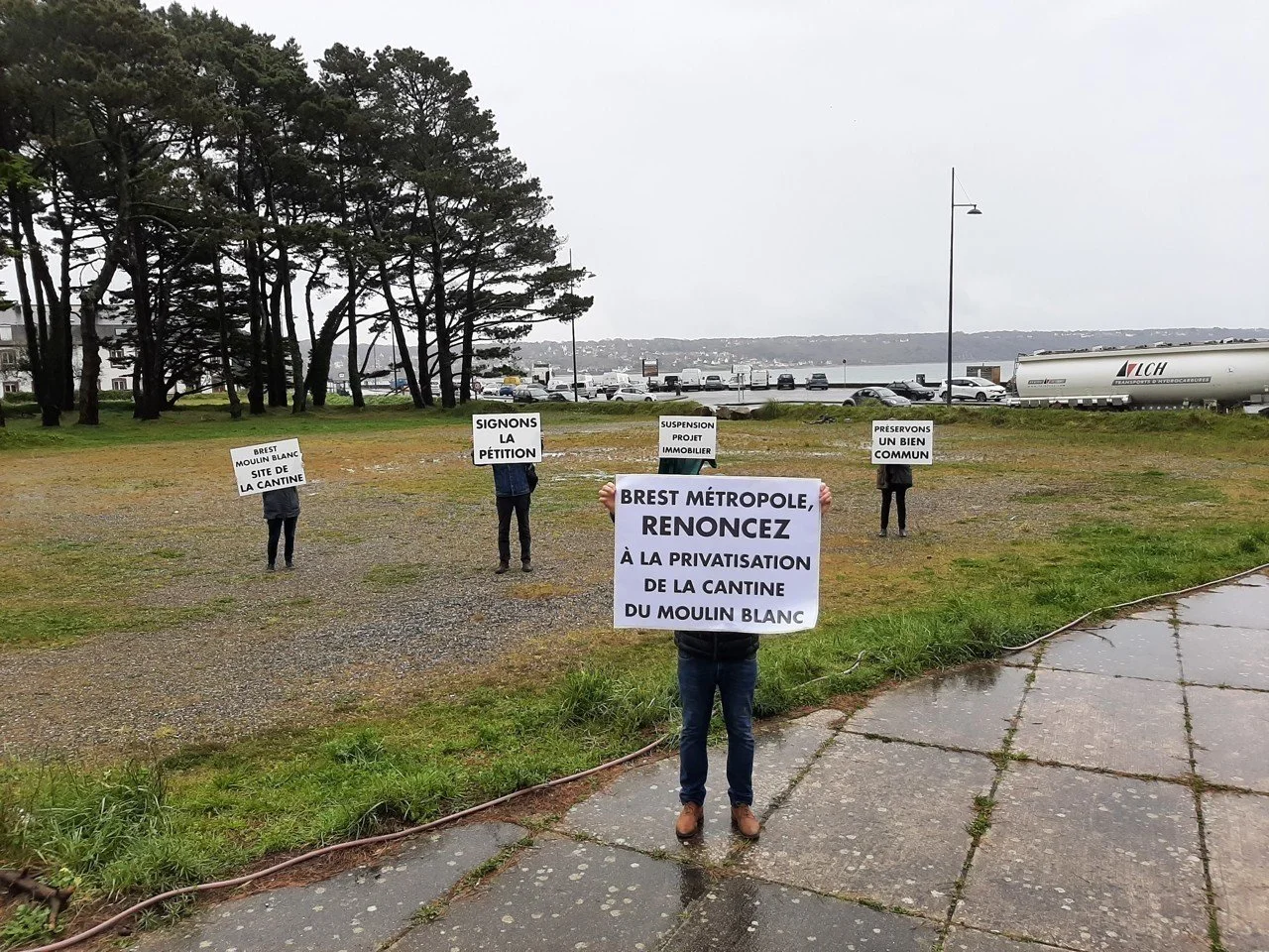 Groupe de personnes tenant des pancartes en protestation contre la privatisation d'une cantine, avec des arbres, un parking et une rivière en arrière-plan.