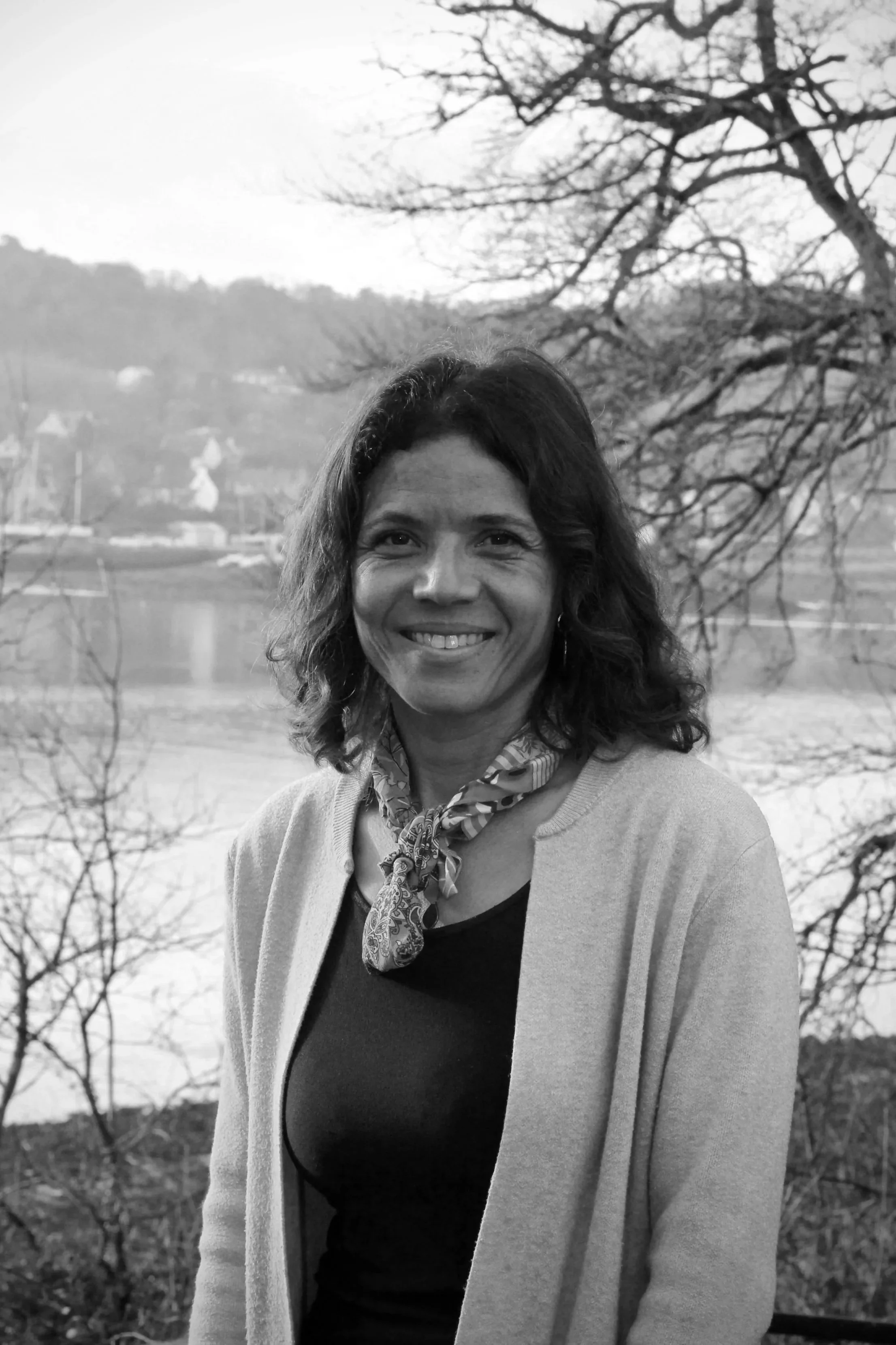 Une femme souriante avec des cheveux courts et bouclés, portant un pull et un foulard, en extérieur près d'un lac ou d'une rivière avec des arbres et des collines en arrière-plan.