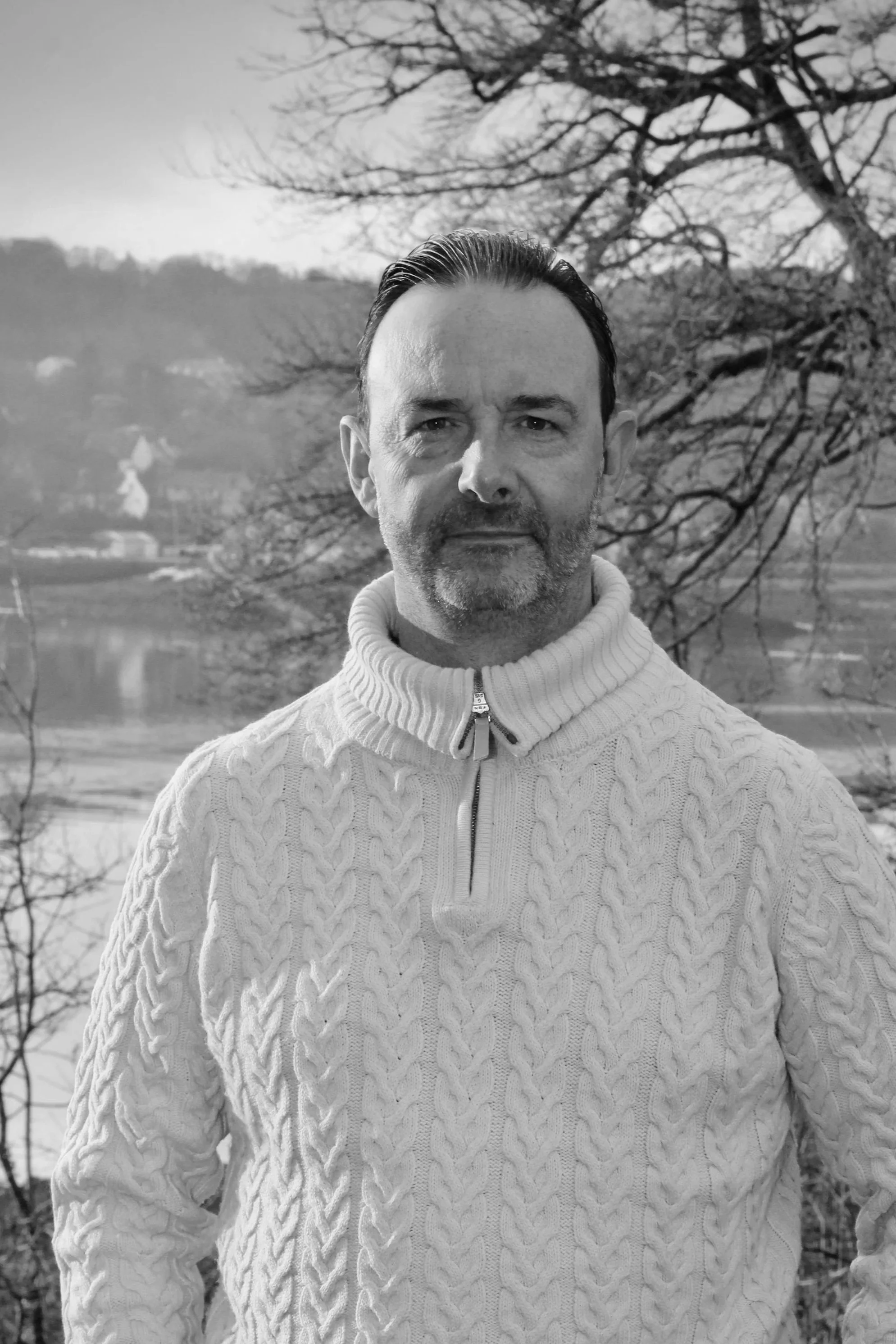 Un homme avec une barbe porte un pull en laine à col roulé, face à un paysage naturel avec des arbres et une rivière en arrière-plan.