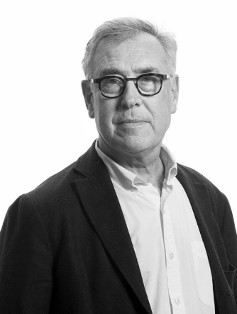 Homme d'âge moyen avec des cheveux gris, portant des lunettes et une chemise blanche, vêtu d'une veste noire, posant à côté d'un fond blanc.