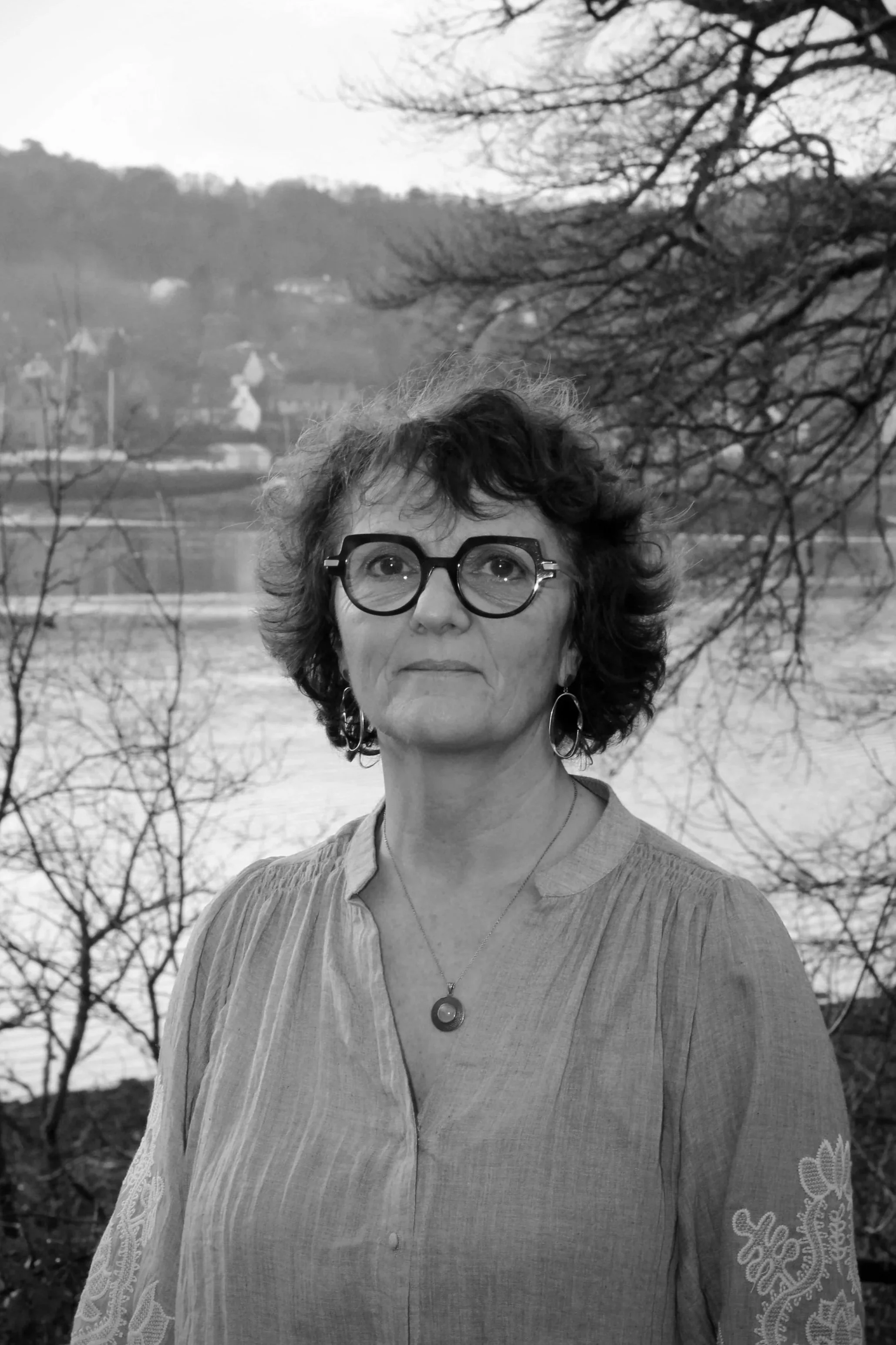Portrait d'une femme portant des lunettes rondes et un collier, avec un paysage en arrière-plan comprenant des arbres sans feuilles et un lac.