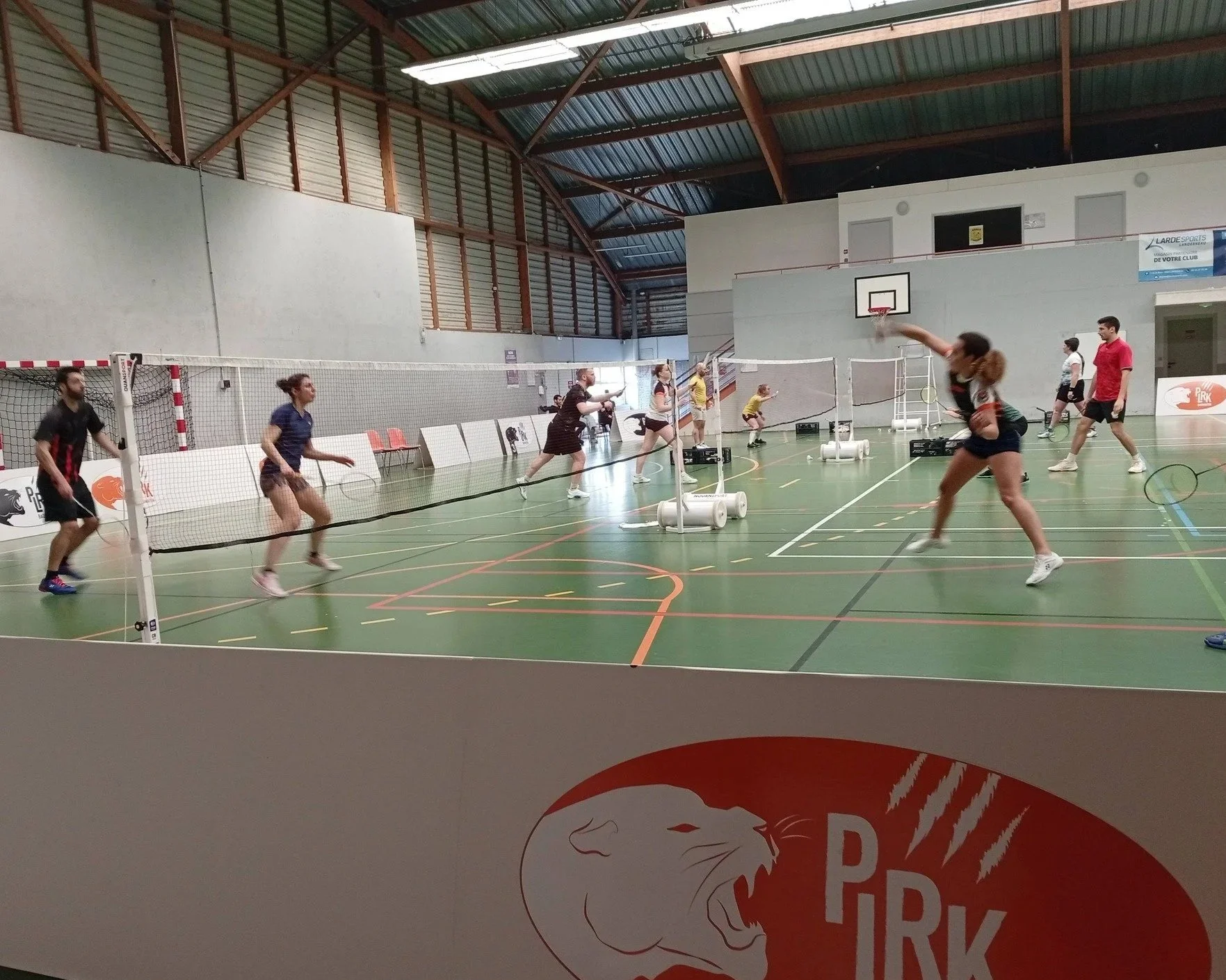Une scène intérieure de badminton où plusieurs personnes jouent au badminton, avec un filet séparant deux camps, dans une salle avec un gros plafond en bois et des murs blancs.