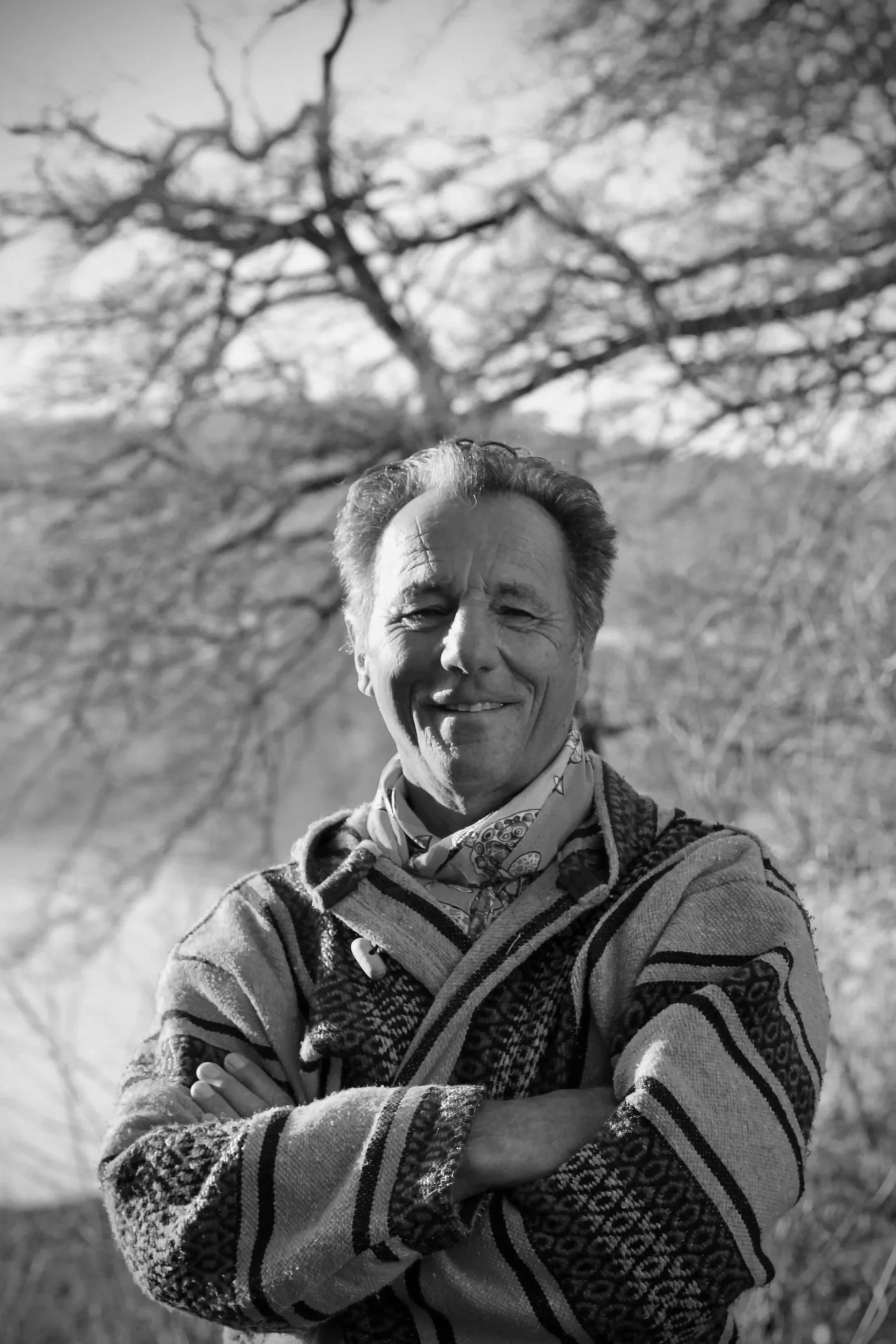 Un homme souriant avec les bras croisés, portant un pull à motifs et une écharpe, se tient devant un arbre nu en arrière-plan.