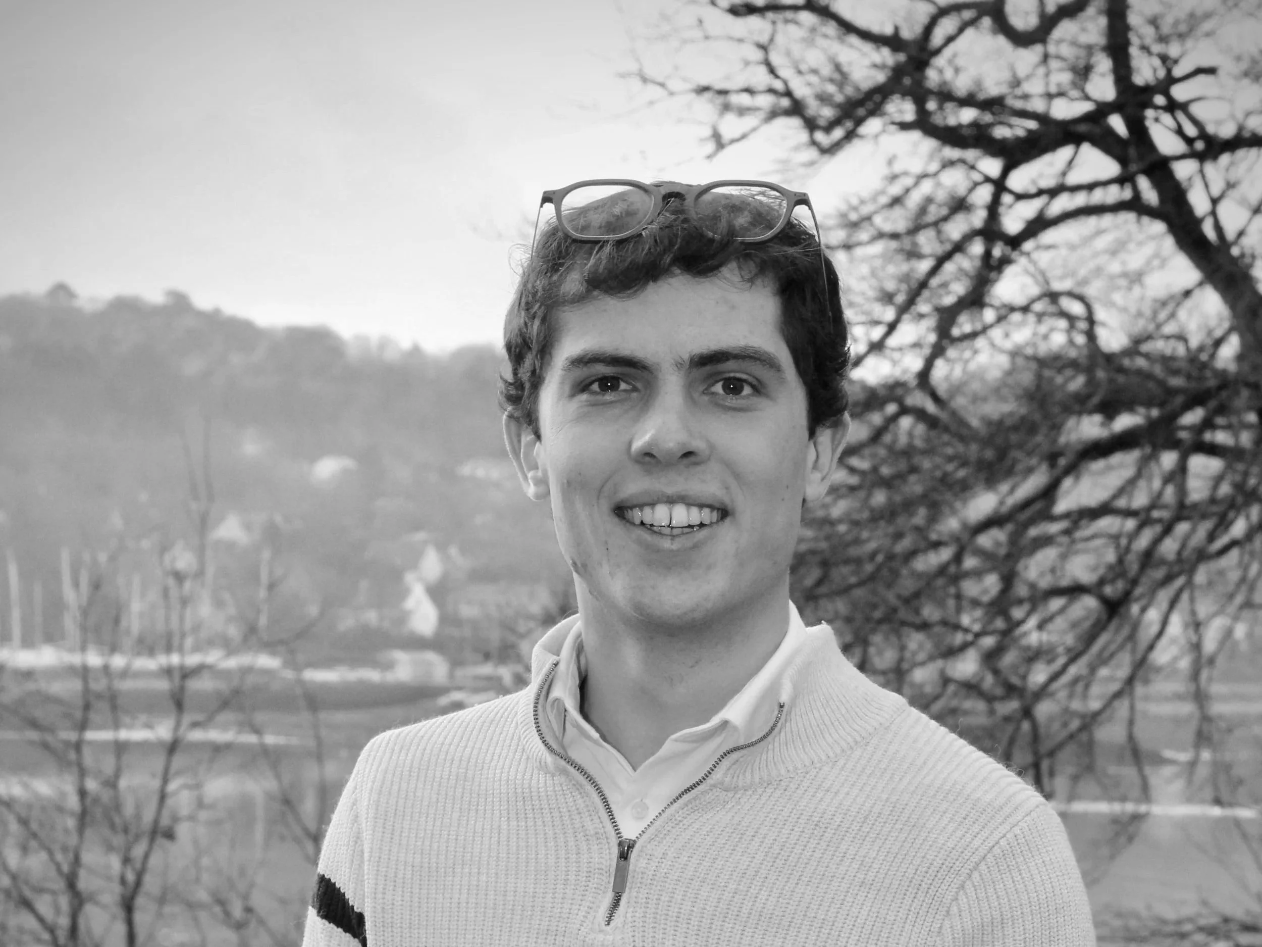 Jeune homme souriant à l'extérieur, portant des lunettes sur la tête et une veste, avec un paysage de nature en arrière-plan.