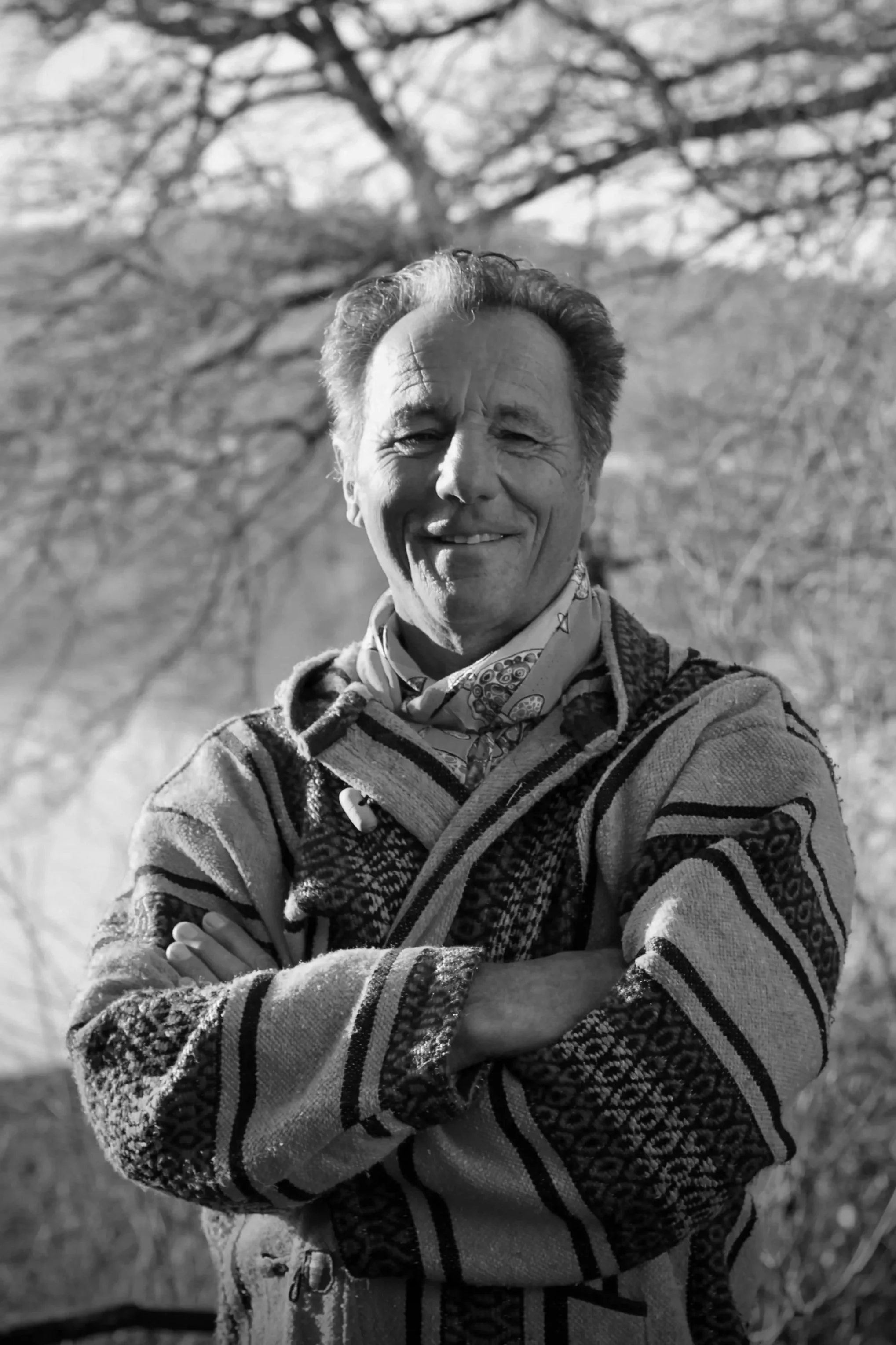 Un homme souriant avec les bras croisés, portant un pull à motifs, une chemise et un foulard, en plein air, avec des arbres en arrière-plan.