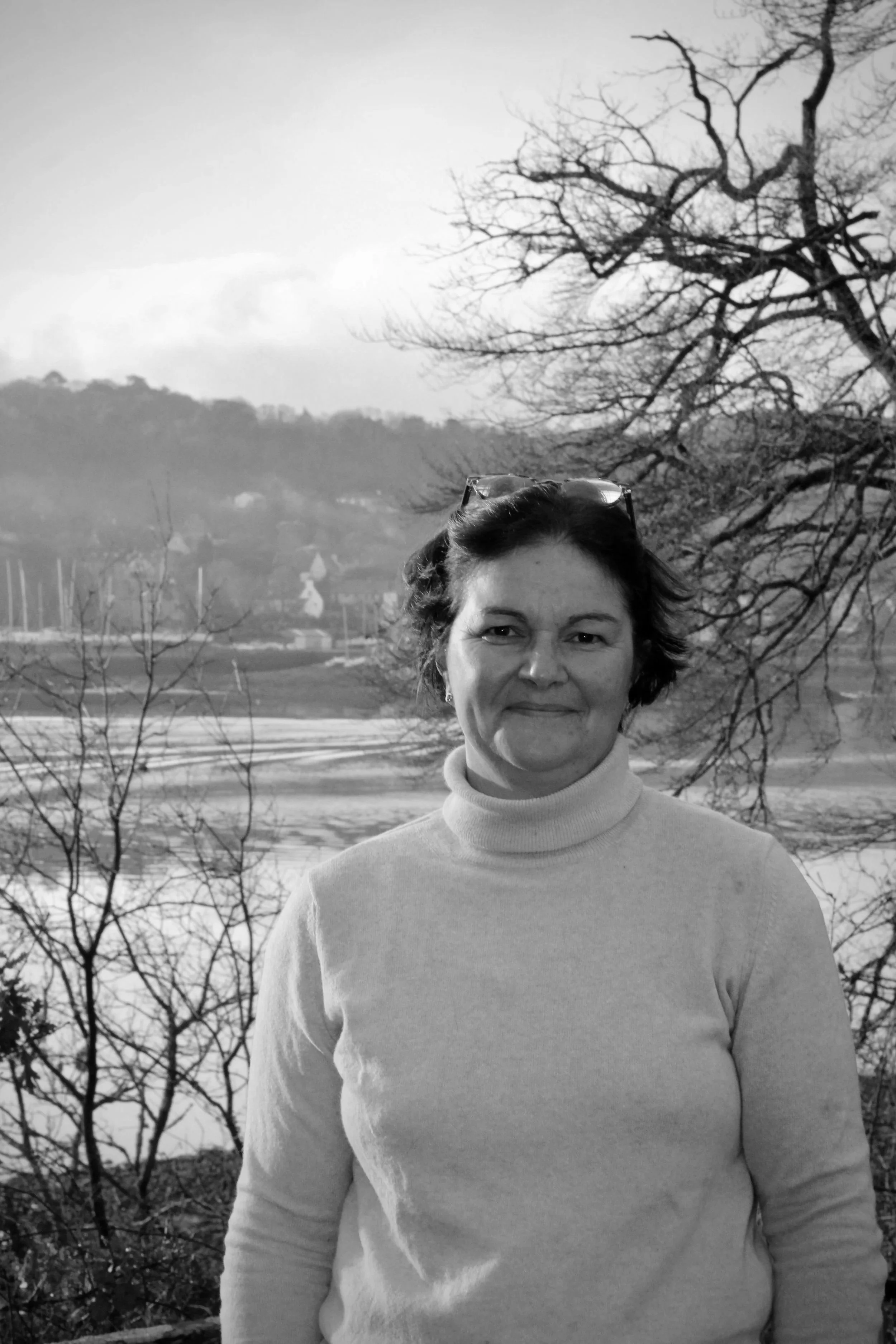 Une femme souriante portant un pull roulé, avec des lunettes sur la tête, devant un paysage hivernal avec des arbres dénudés et un lac en arrière-plan.