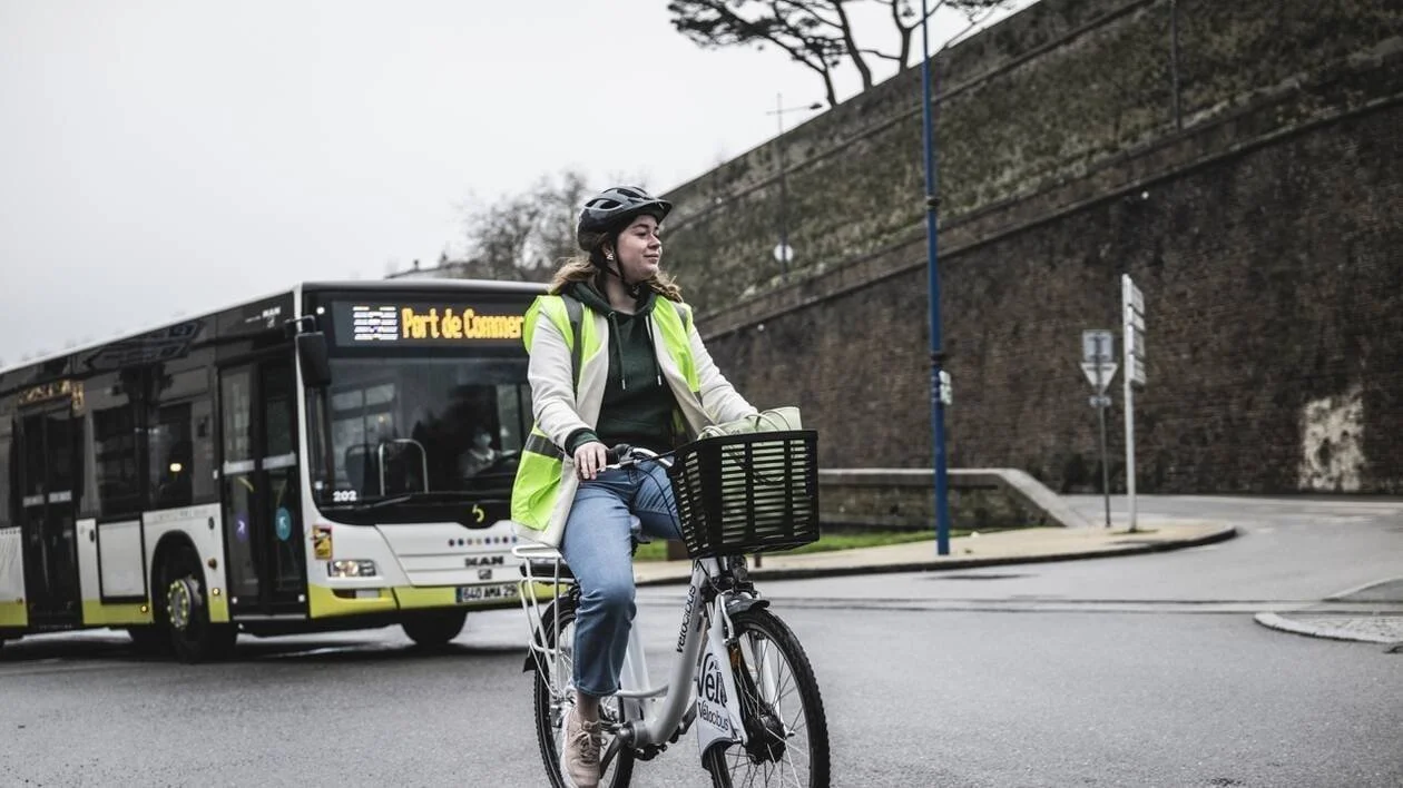 Une femme portant un casque et un gilet jaune à la bicyclette, avec un bus derrière elle sur une route citadine.