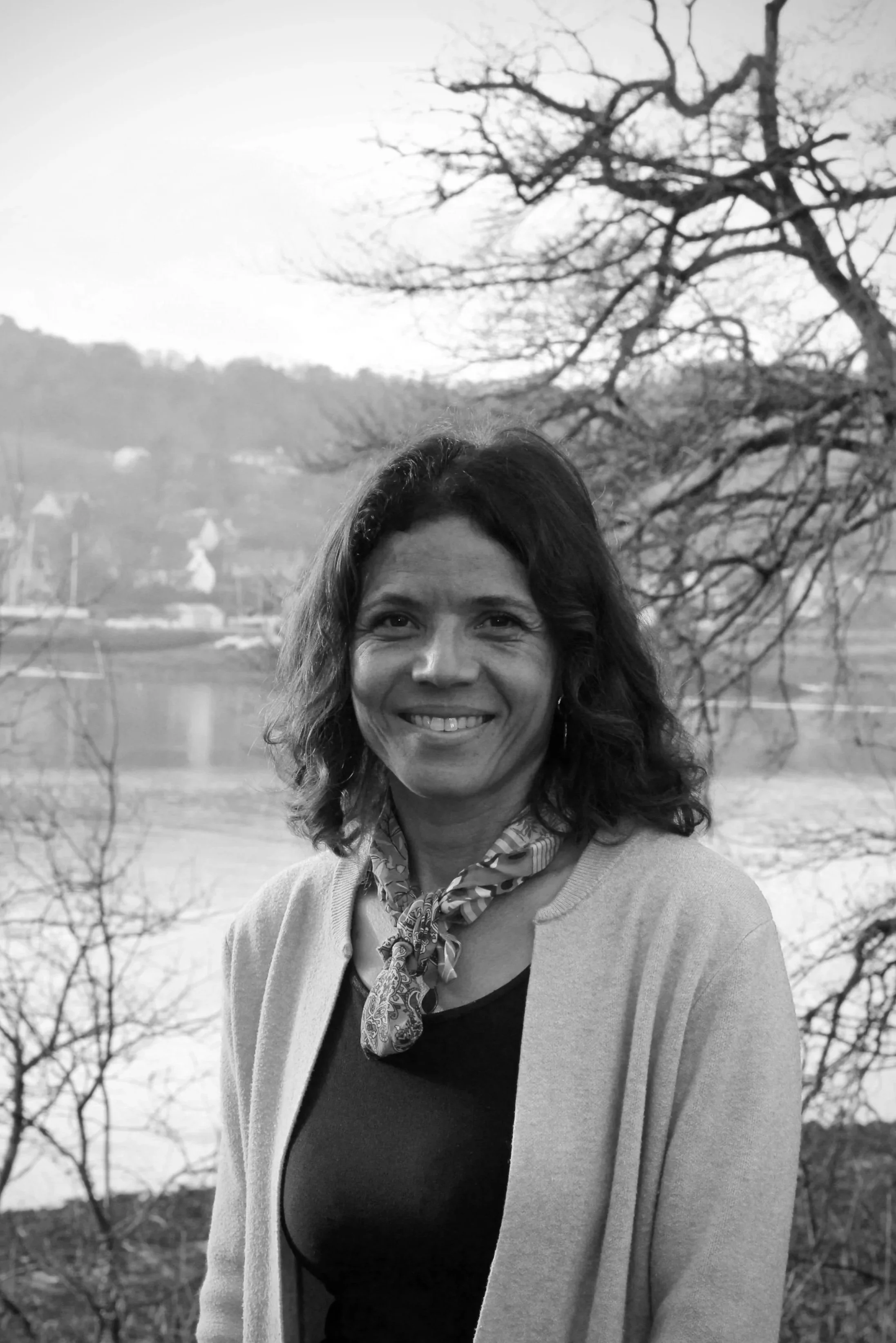 Une femme souriante avec des cheveux bruns bouclés, portant un foulard, un t-shirt noir et un cardigan, se tient près d'un plan d'eau avec des arbres et une colline en arrière-plan, en noir et blanc.