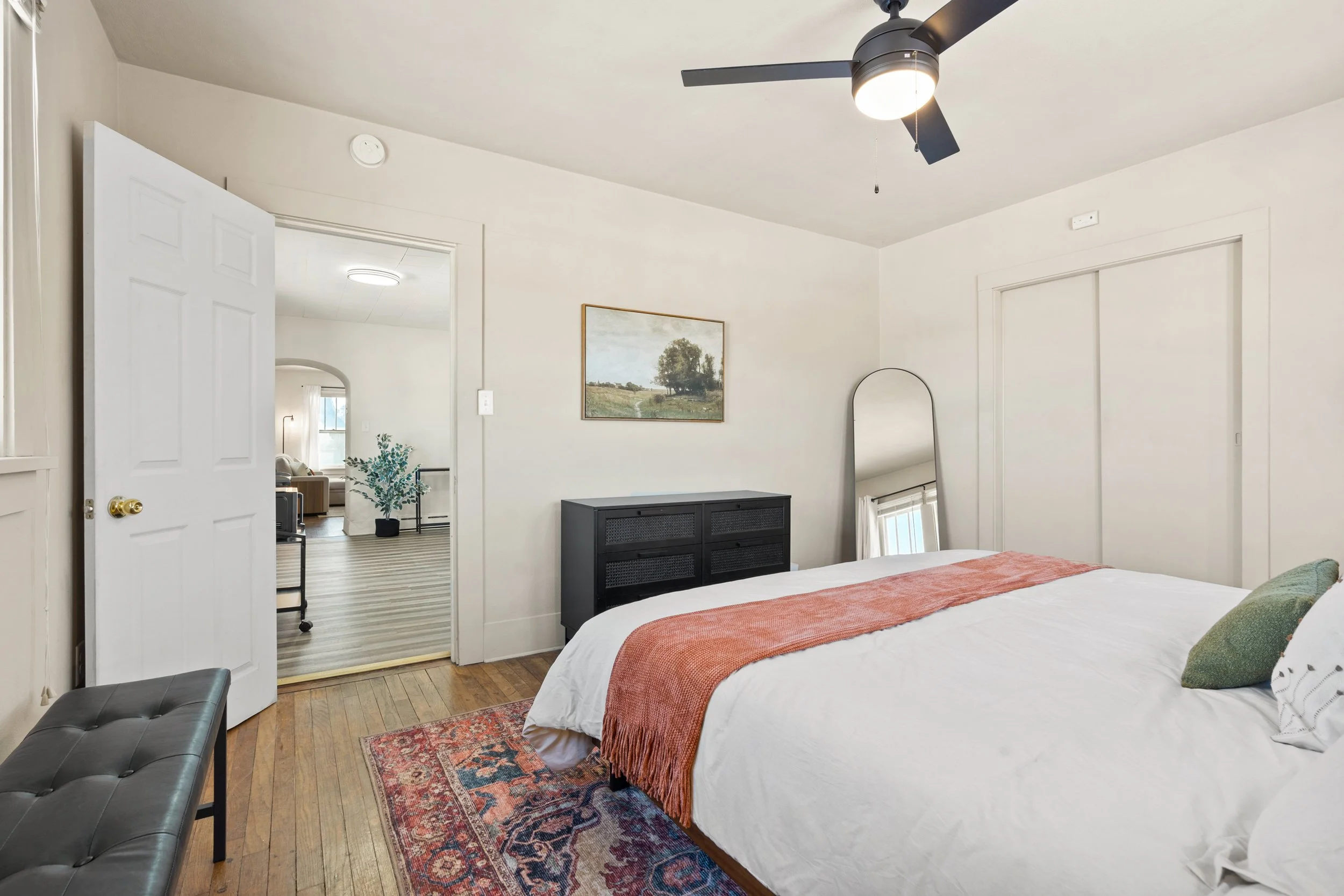 A bedroom with a bed, decorative pillows, and a burnt orange throw blanket. A black dresser, a floor mirror, and a portrait painting of a landscape with trees hanging on the wall. An open door leads to a room with a potted plant and a sitting area. A ceiling fan with a light is installed on the ceiling.