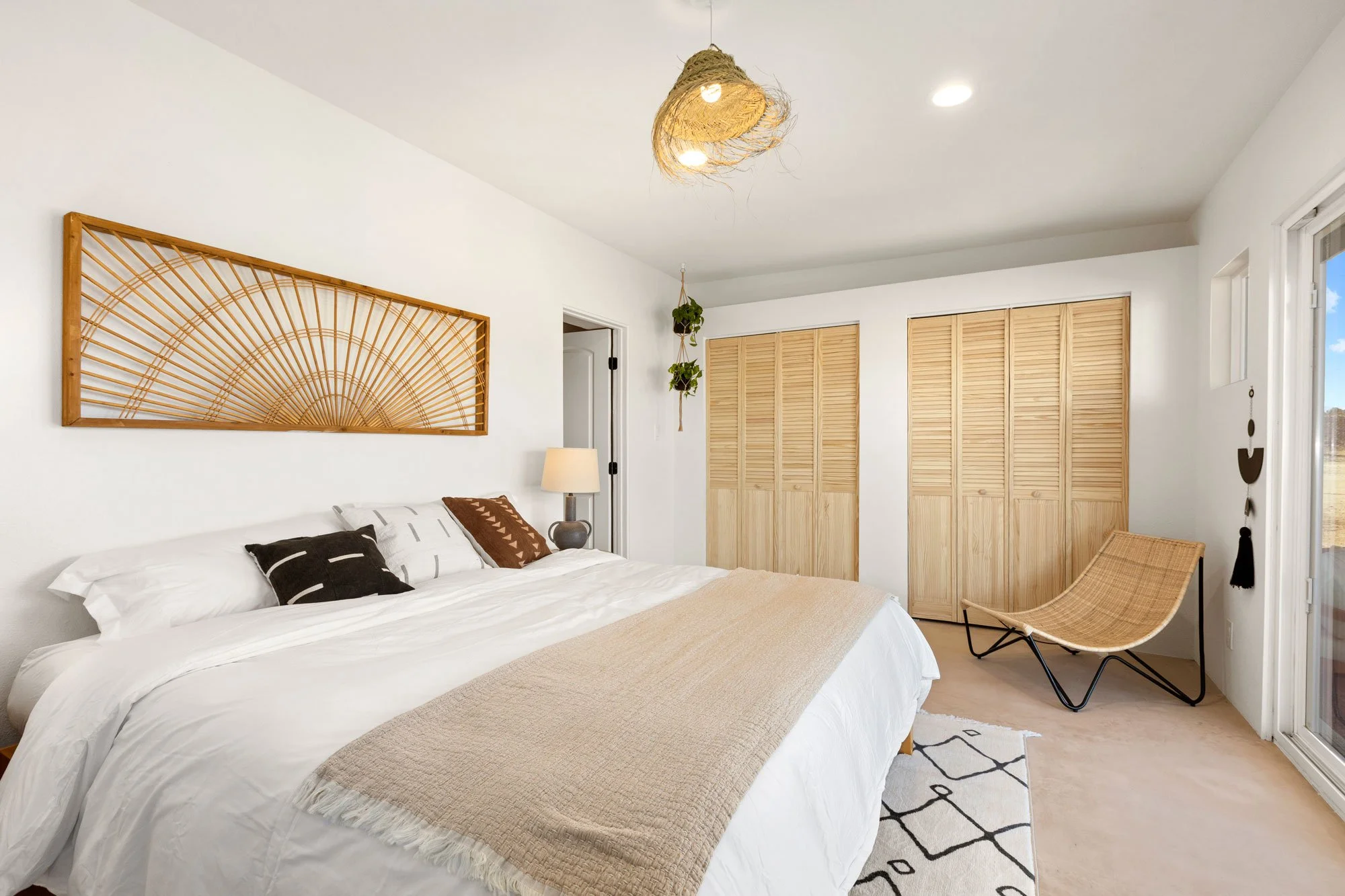 A bedroom featuring a bed with white bedding and black, white, and brown pillows, a bedside lamp, a hanging plant, wooden closet doors, a rattan lounge chair, a drawstring rug, and a decorative wall piece with a sunburst design.