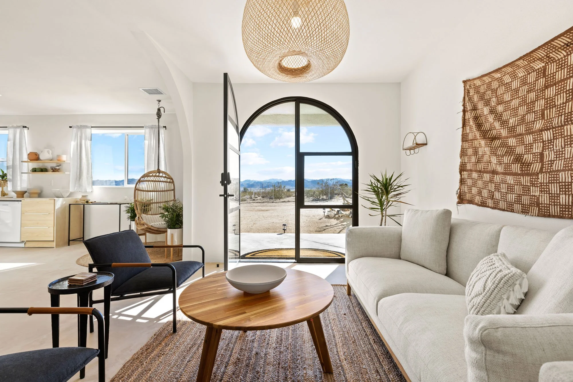 Bright living room with arched glass door opening to desert landscape, white couch, black and brown chairs, round wooden coffee table, hanging wicker chair, potted plant, woven wall hanging, minimalist decor