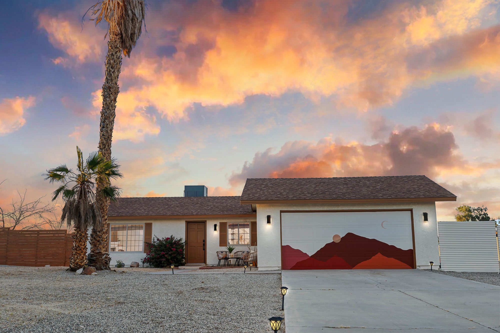 A single-story house with a mountain mural on the garage door, a gravel yard, two palm trees, a sunset sky with pink and orange clouds, and outdoor lighting.