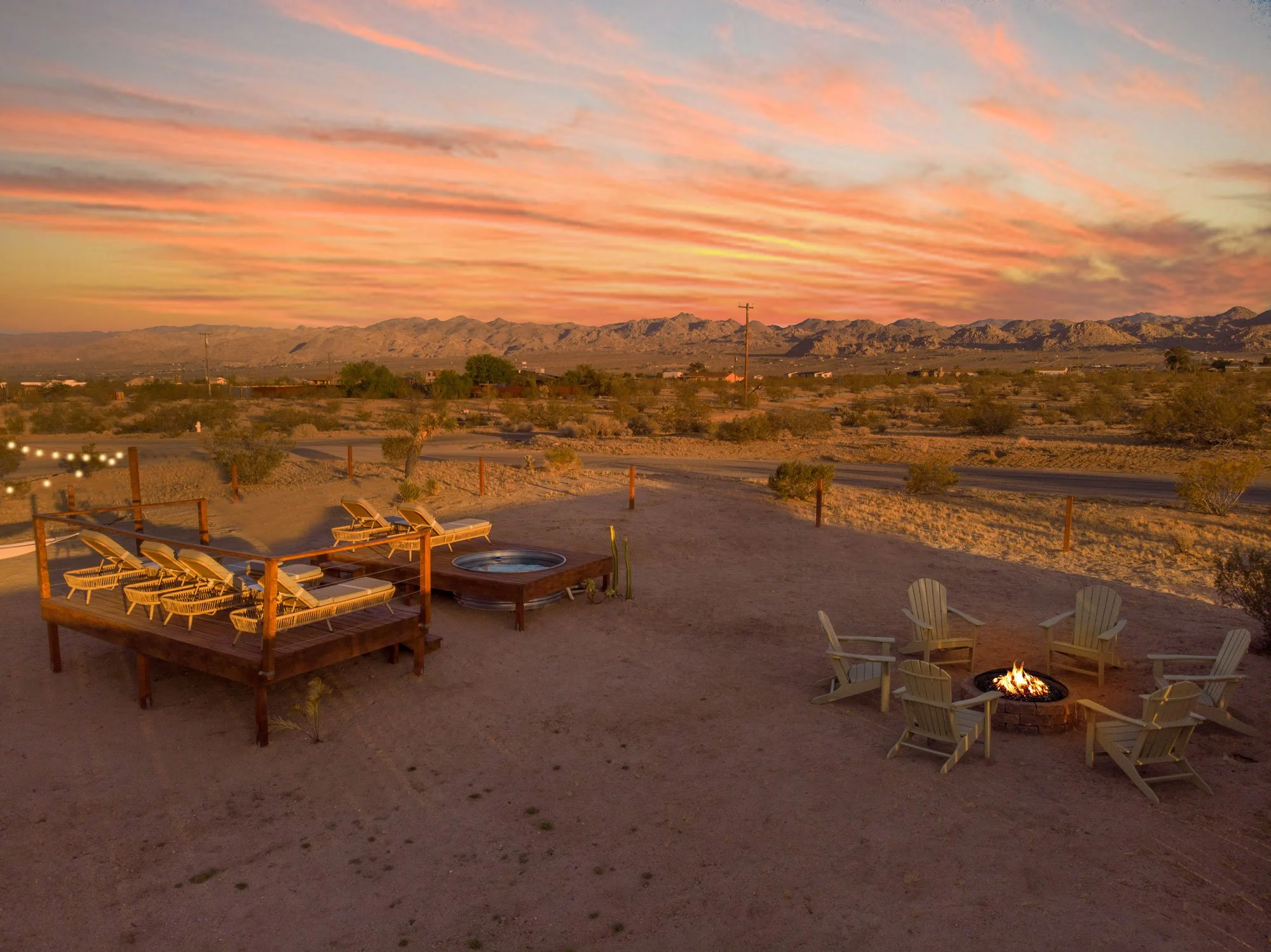 Desert landscape at sunset with mountains in the background. In the foreground, there are outdoor lounge chairs, a small hot tub, and a circle of Adirondack chairs around a fire pit.