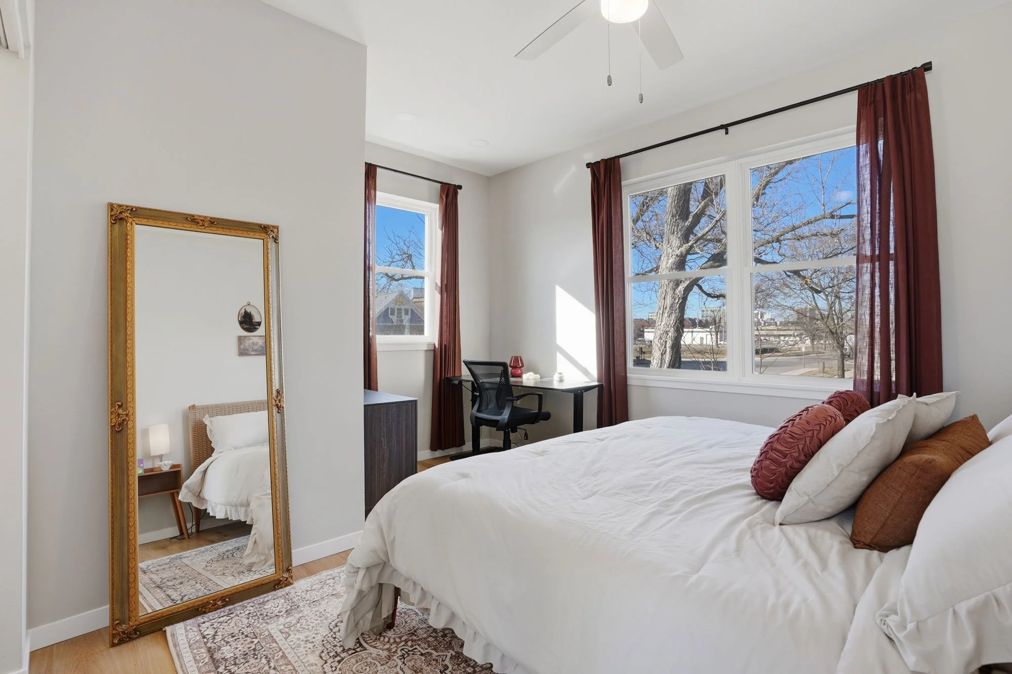 Bright bedroom with large windows, white walls, and a white bed with brown and cream pillows. A tall ornate mirror leans against the wall, and a small desk with a black chair and red lamp is placed by the windows. The room has hardwood floors and a ceiling fan.