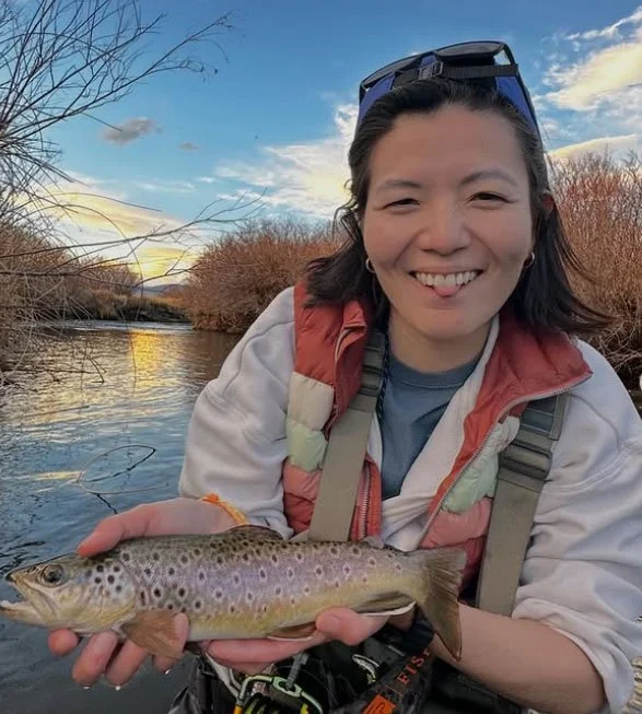Smiling woman holding a small fish outdoors near a river during sunset, with trees and a partly cloudy sky in the background.