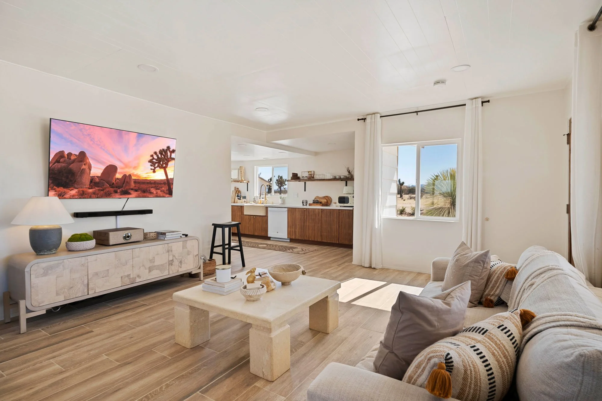 Living room with a beige couch and throw pillows facing a large flat-screen TV mounted on a white wall. The TV displays a desert landscape with rocks and a Joshua tree at sunset. There is a white console table beneath the TV, with decorative items and books. To the right, a window with white curtains lets in natural light, showing a desert scene outside. The room has wooden flooring and an open kitchen with wooden cabinets and shelves in the background.