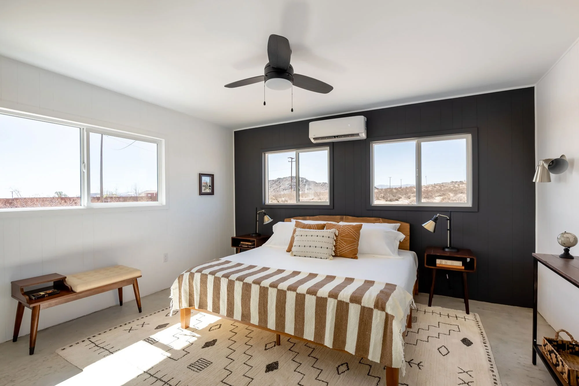 Bedroom with white walls, black accent wall behind bed, large windows showing desert landscape, wooden bed with striped blanket, pillows, black bedside tables with lamps, ceiling fan, and air conditioner.