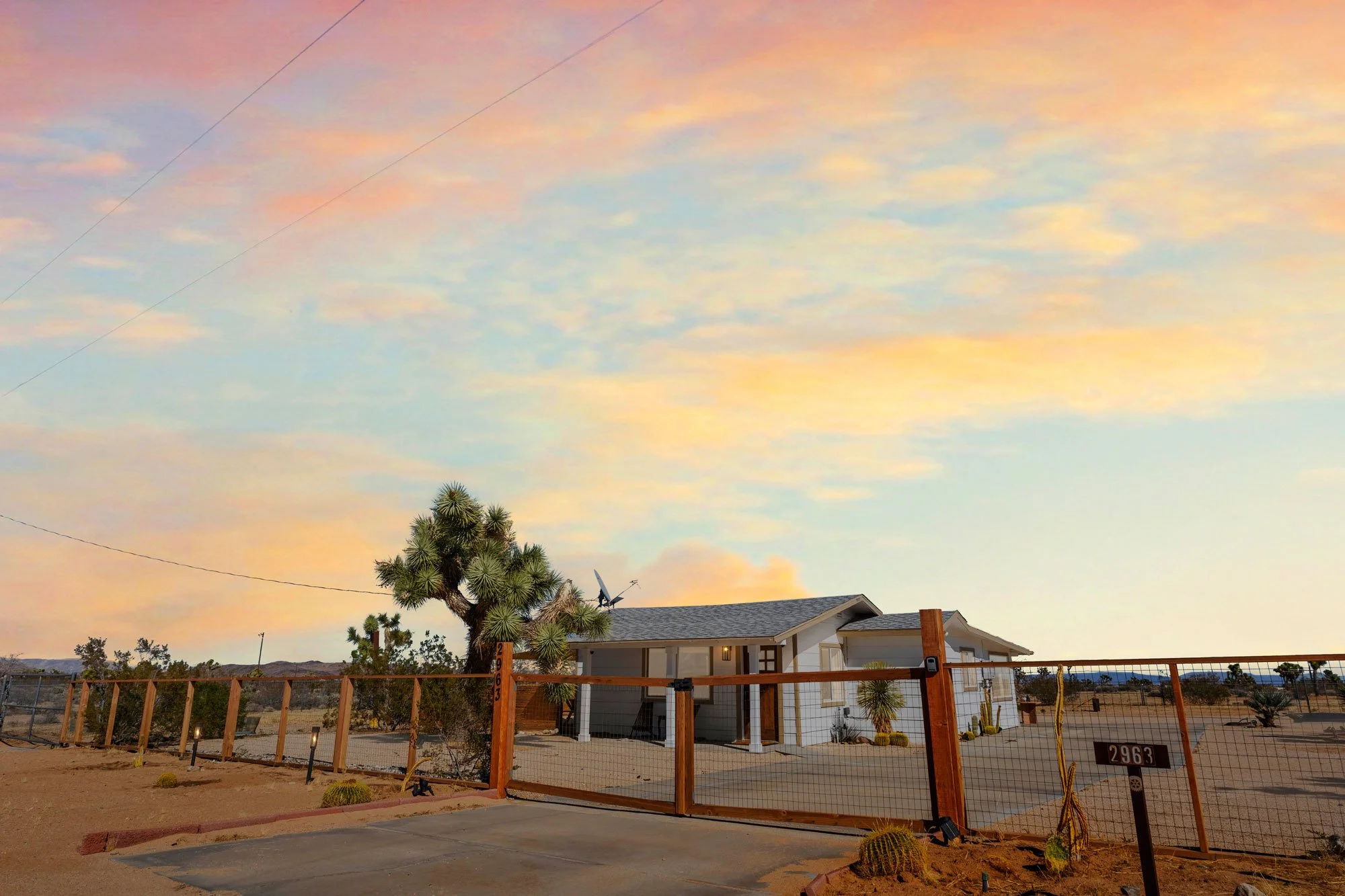 A house in a desert landscape with a cactus in the front yard, a gate, and a sky with pink and orange clouds at sunset.