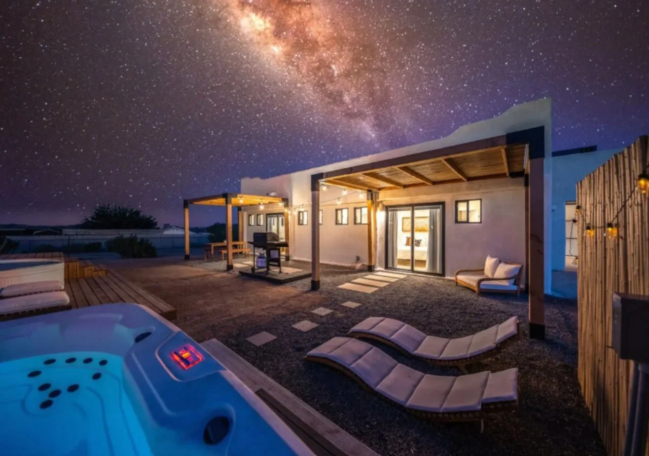 Nighttime view of a modern house with outdoor lounge chairs, a hot tub, and string lights under a starry sky with the Milky Way galaxy visible.