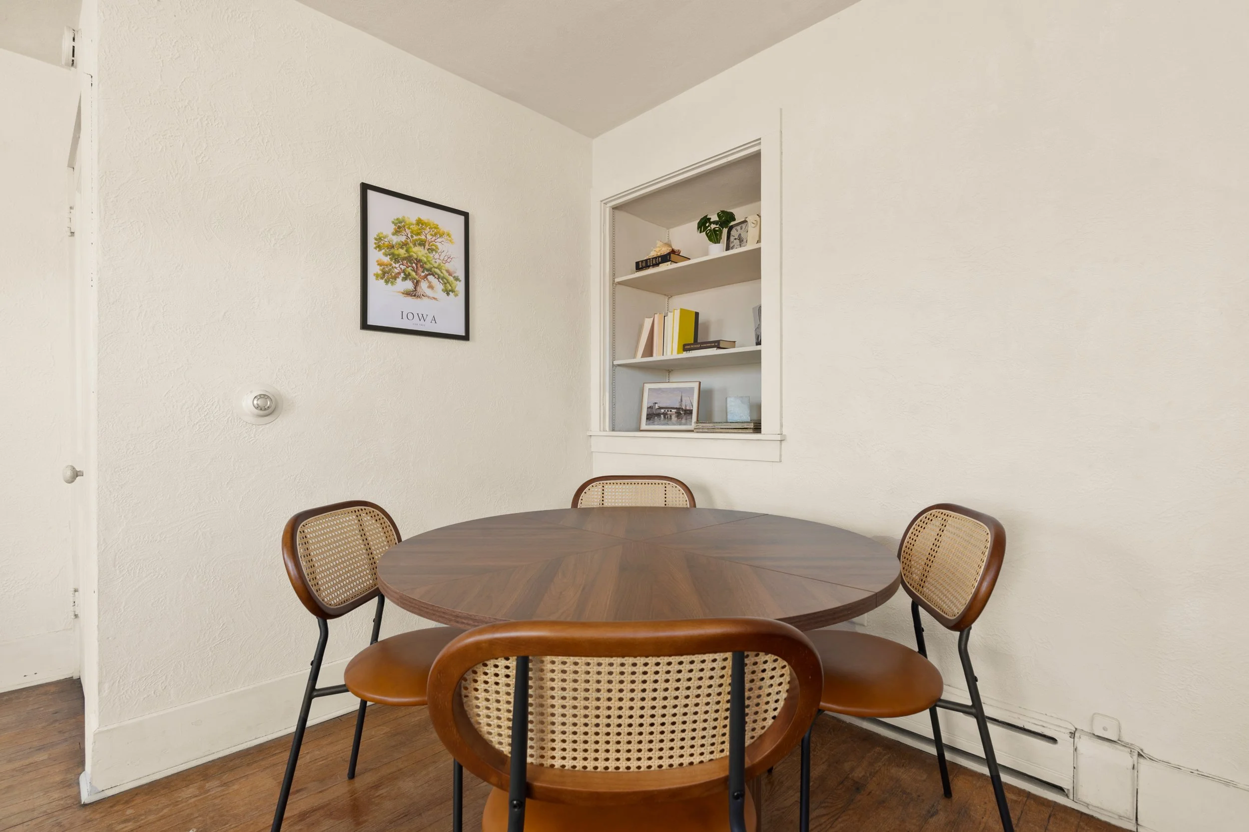A round wooden dining table with four matching chairs in a room with white textured walls, a framed tree poster labeled "Iowa," and built-in shelves with books and framed photos.