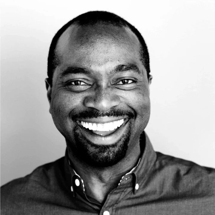 A smiling man with short hair, a beard, and mustache, wearing a button-up shirt, standing against a plain background.