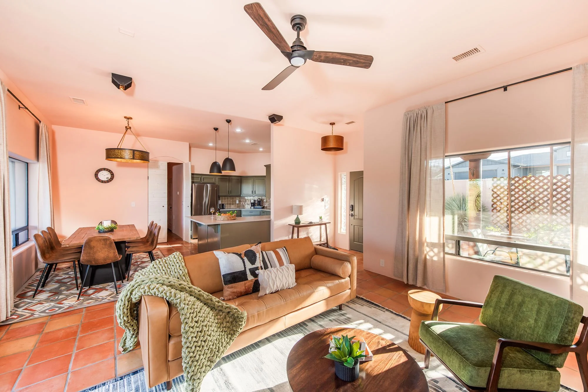 Spacious living room with tan sofa, green armchair, and wooden coffee table, adjacent to dining area with wooden table and six chairs, open kitchen with gray cabinets, and large window with curtains letting in sunlight.
