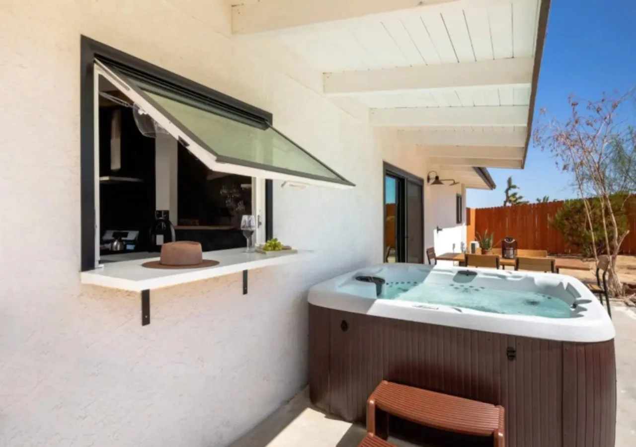 Outdoor patio with hot tub, table and chairs, and an open window showing a kitchen inside. Bright sunny day, with a tree and wooden fence in the background.