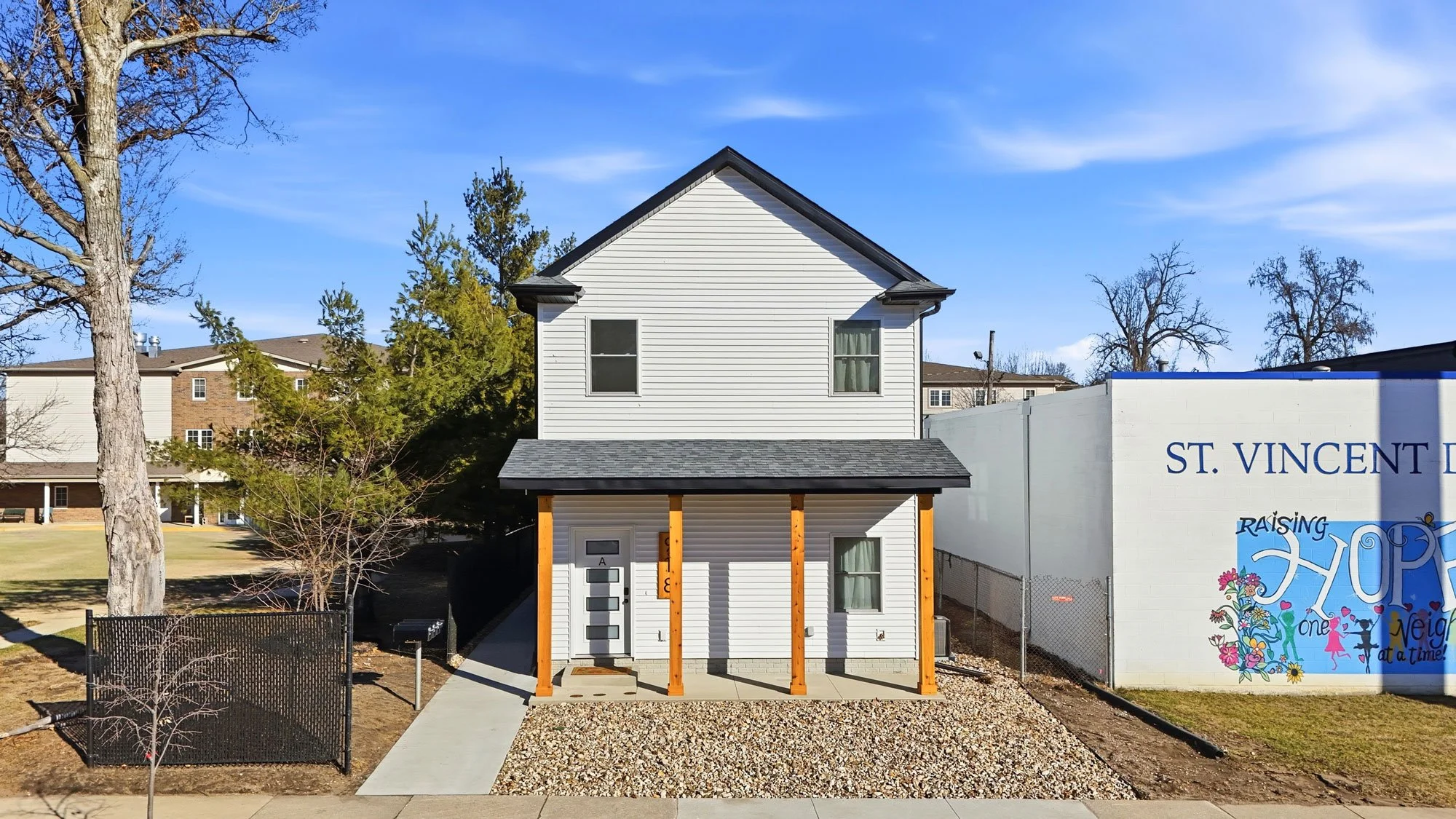 A white, two-story house with a small covered porch supported by four wooden posts. The house has two windows on the upper floor and three on the lower floor, with a modern front door. There is a gravel yard in front, a concrete walkway, and a black fence on the left side. To the right, a large white wall features a colorful mural with the word "HOPE" and other decorative elements.