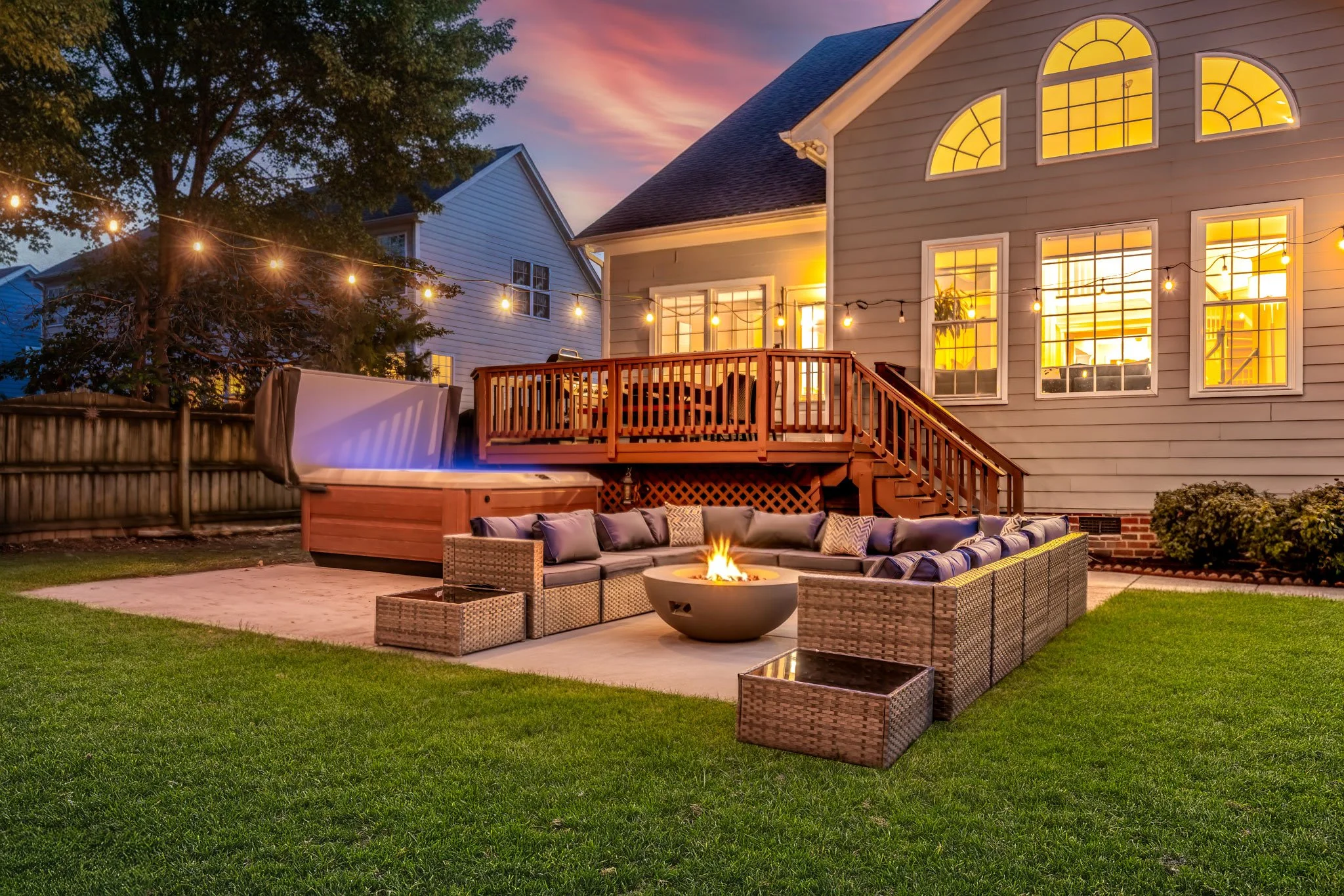 Backyard patio with fire pit, cushioned sectional seating, outdoor hot tub, and string lights at dusk